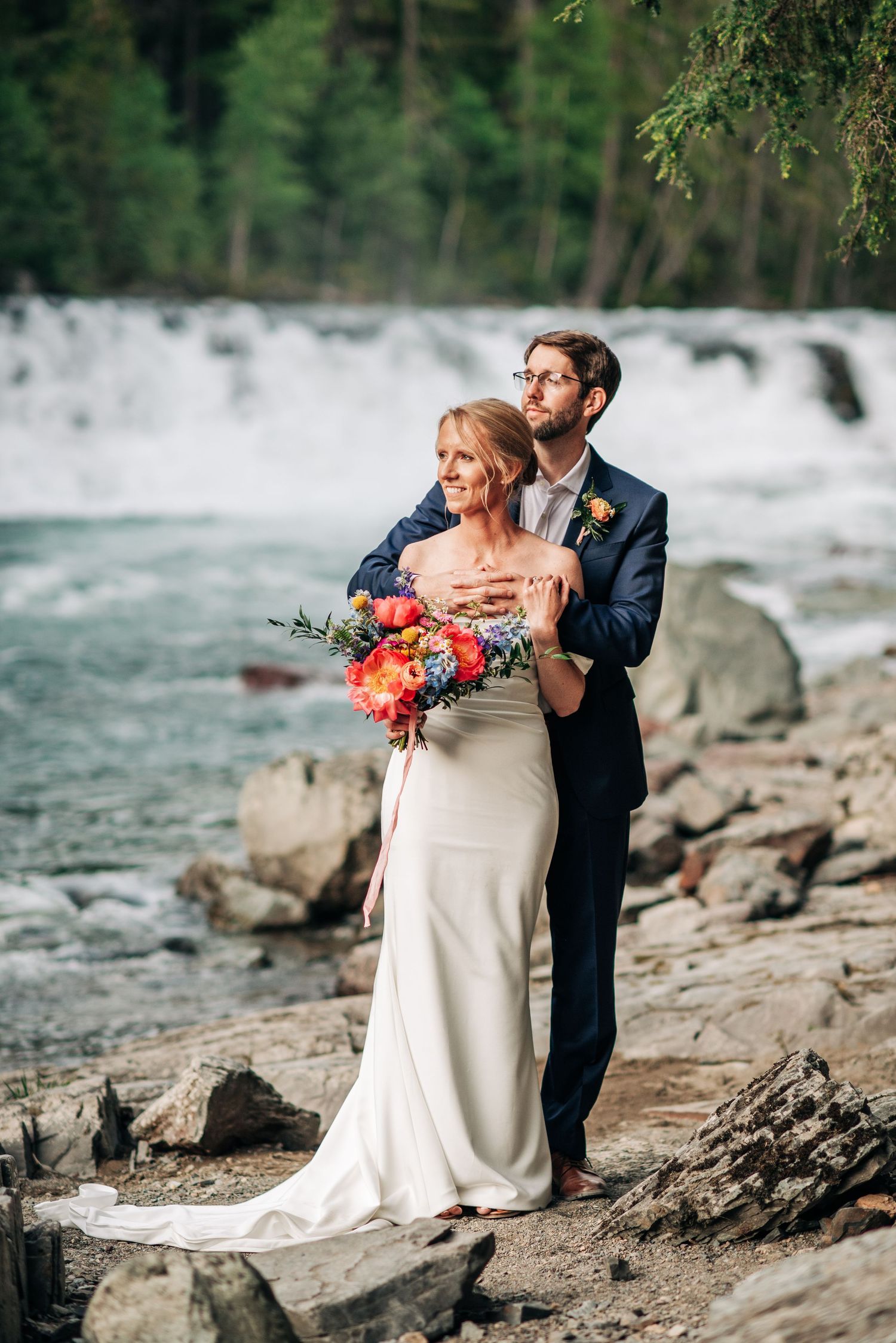 A couple embraces near a rushing waterfall with a colorful bouquet on their wedding day.