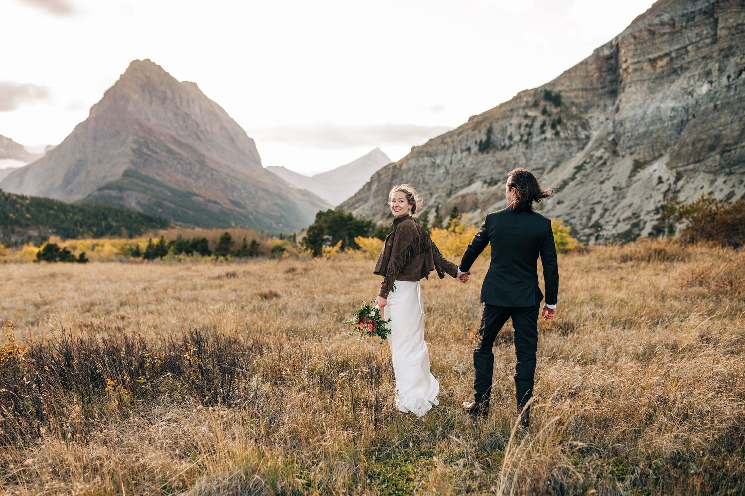 Two people walk hand in hand through golden mountain meadows at sunset in Waterton Lakes National Park.