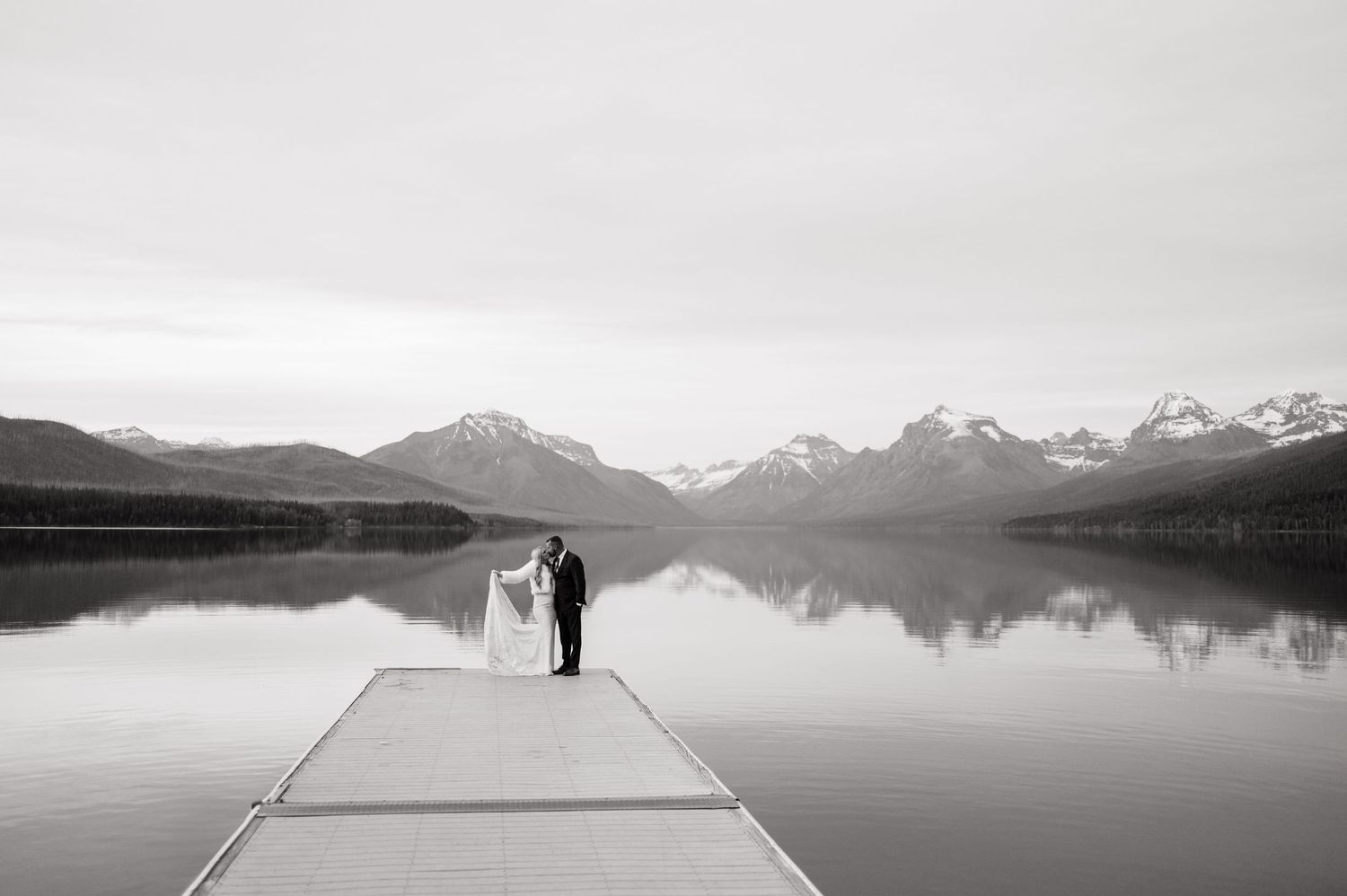 Black and white wedding photo of bride and groom kissing on a dock overlooking a serene mountain lake.