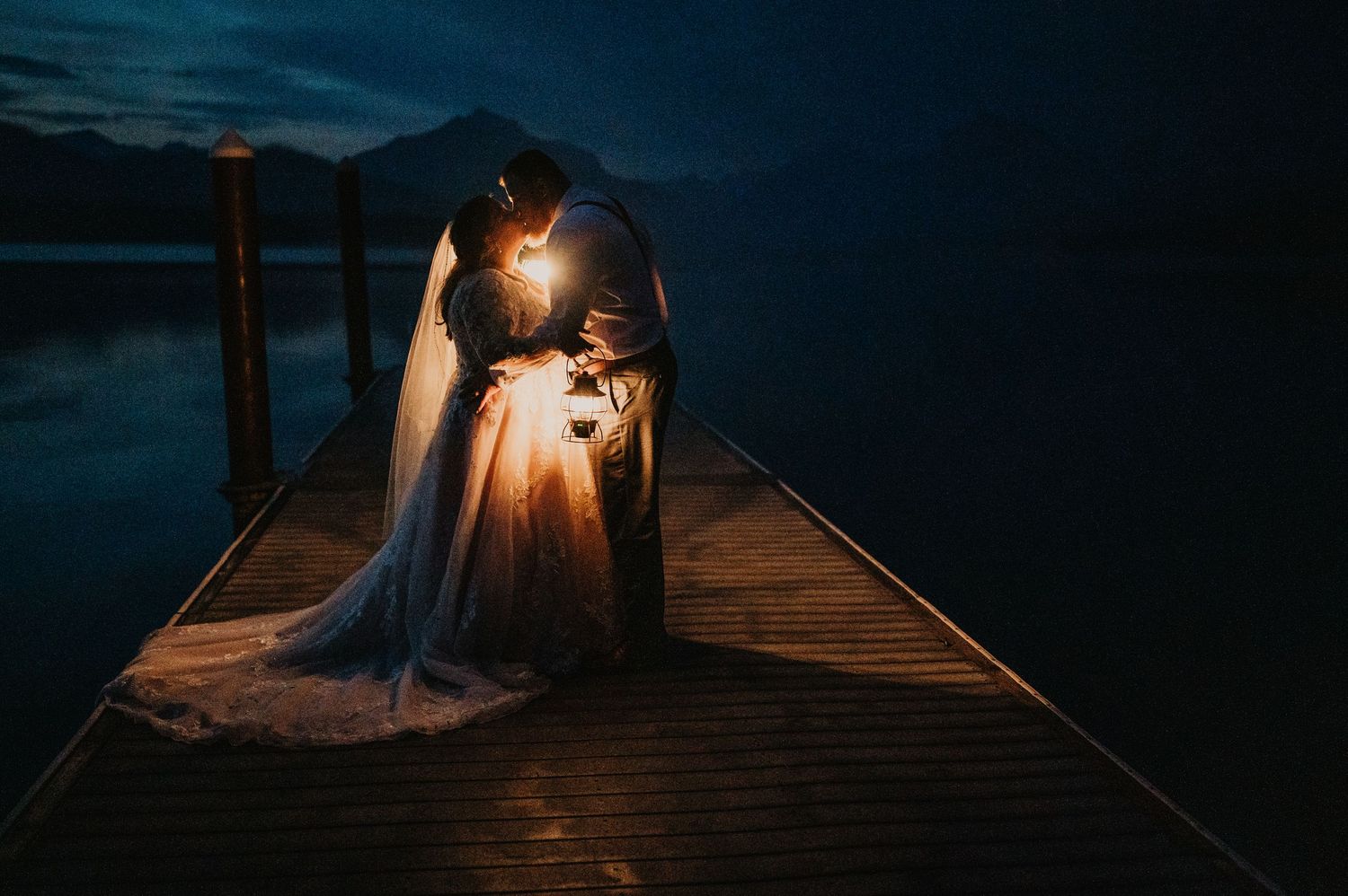 Bride and groom kissing on a wooden dock at night with glowing light, mountains and lake in the background.