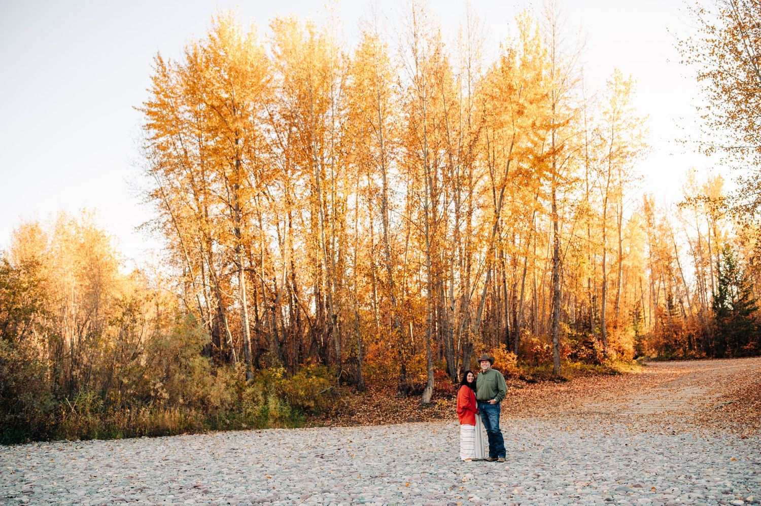 Two people stand together on a gravel path surrounded by golden autumn trees glowing in warm sunlight.
