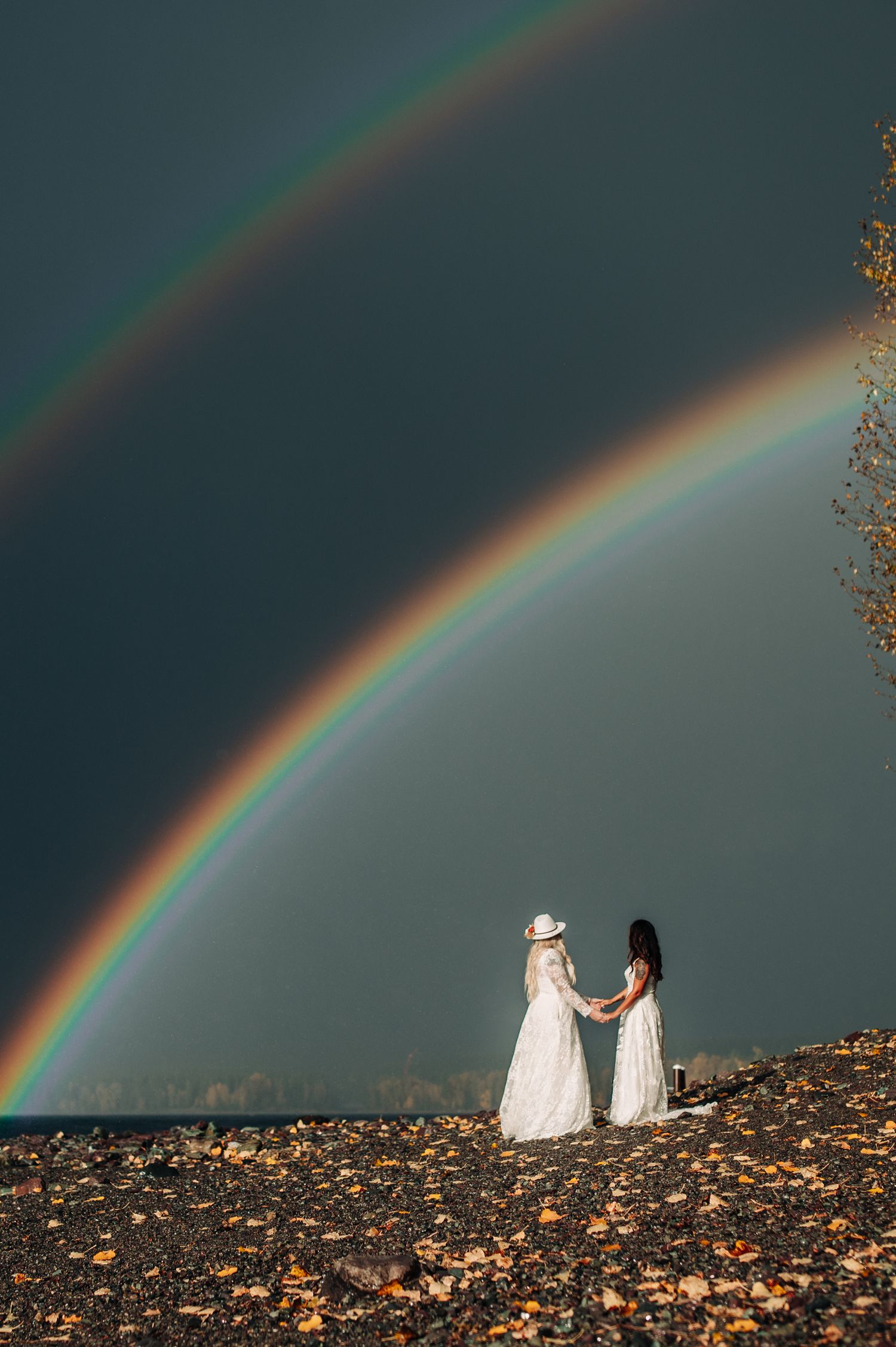 Two figures in white dresses stand beneath a vibrant double rainbow against a stormy sky.