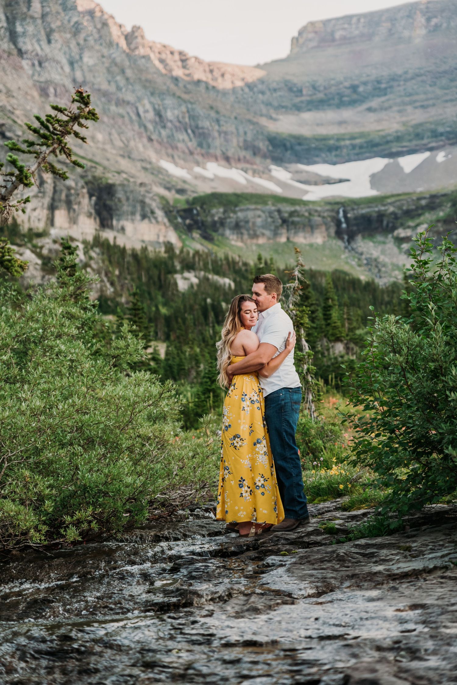 A couple embraces on a mountain trail with dramatic cliffs and snow patches visible in the background.