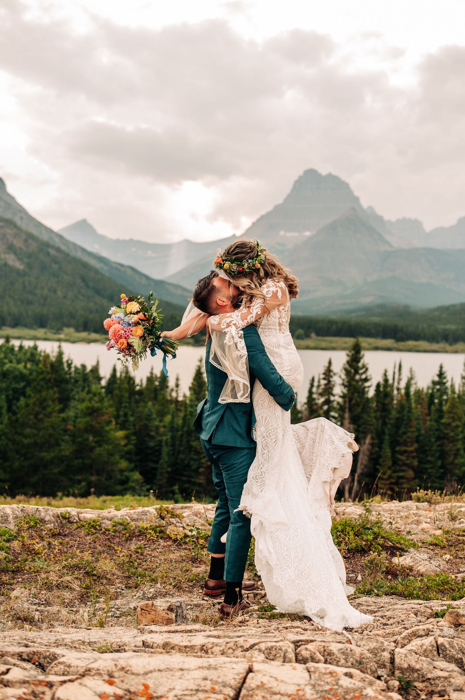 Couple embracing on rocky mountain overlook with scenic lake and peaks in Glacier National Park at sunset.