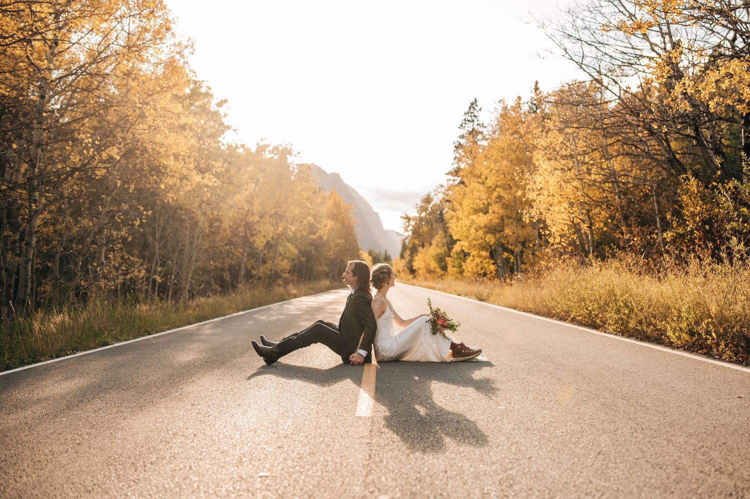 Two people sit on empty autumn road surrounded by golden trees during sunset, creating dreamy warm atmosphere.