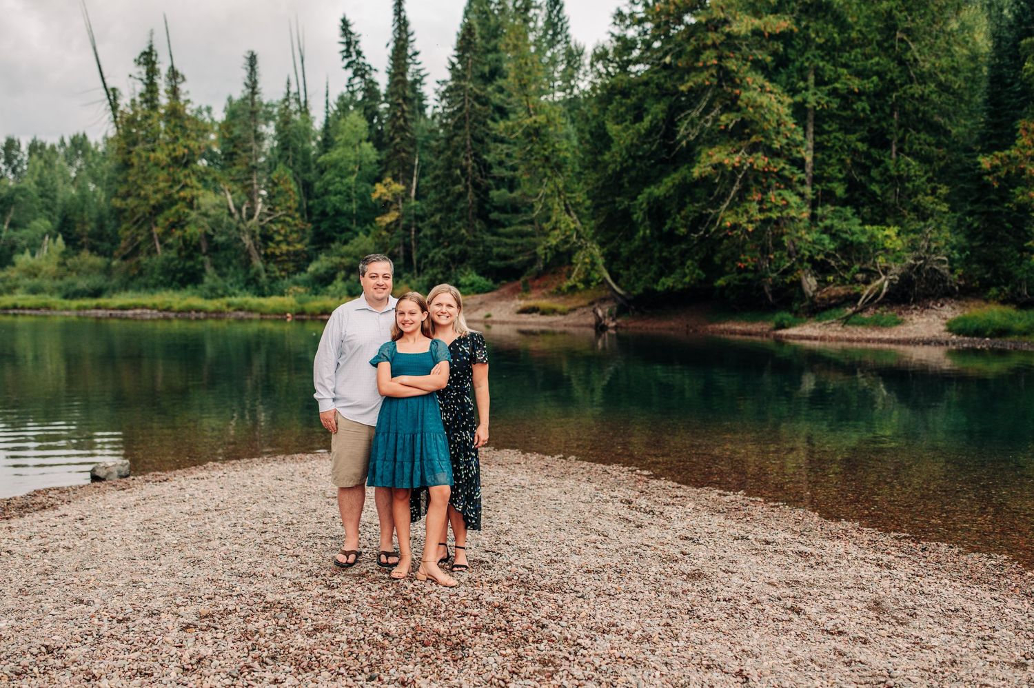Three people stand together on a rocky beach beside a serene lake surrounded by evergreen forest.