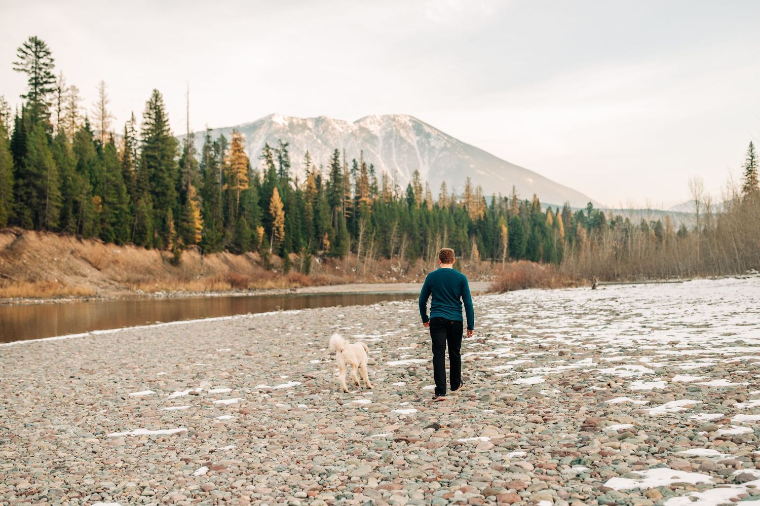 A figure in teal jacket walks with a dog along a snowy riverbank with evergreen forest and mountain backdrop.