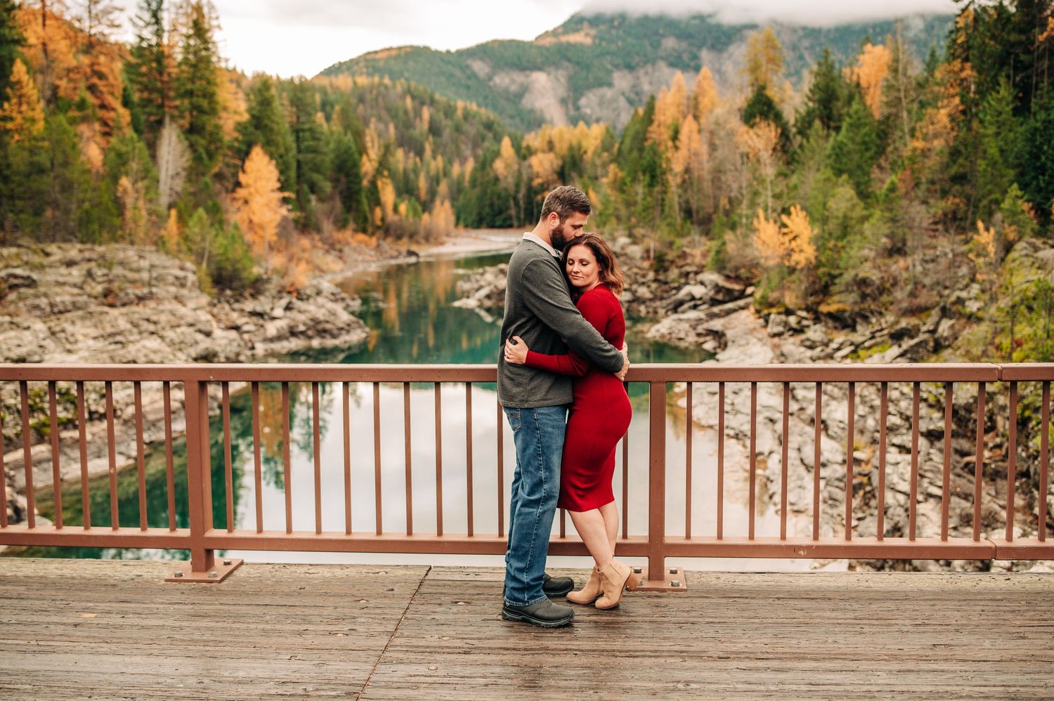 A couple embraces on a wooden bridge overlooking a serene lake surrounded by autumn-colored forest in the mountains.