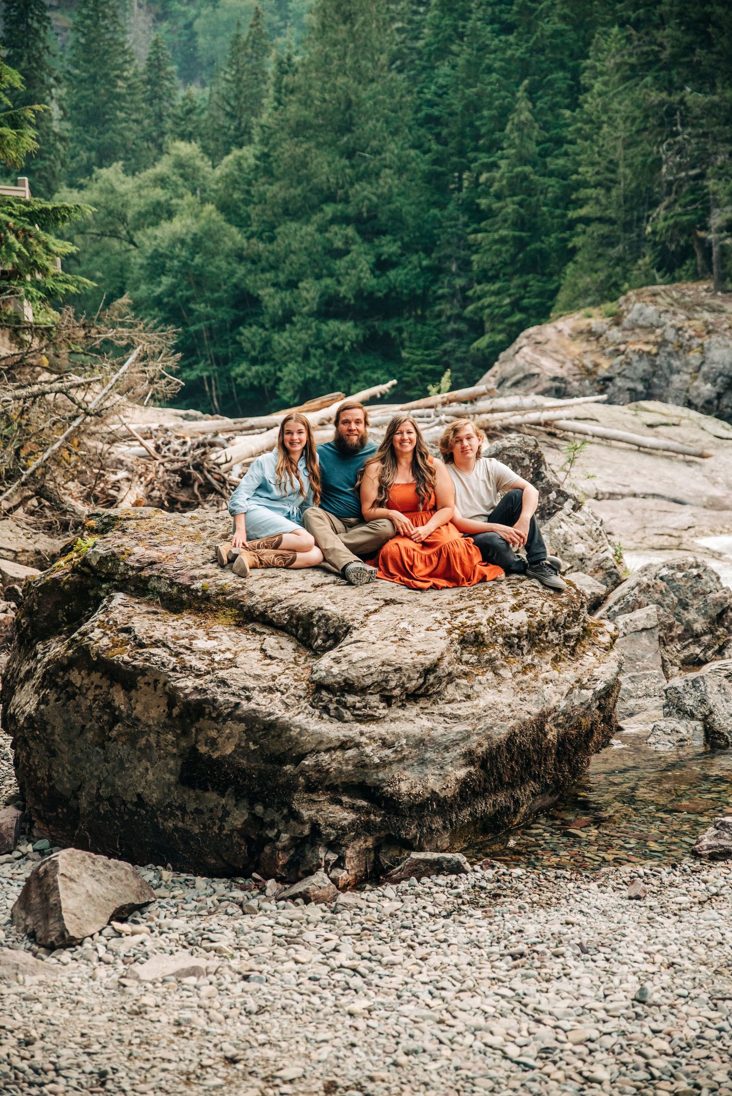 Group relaxes on rocky beach ledge surrounded by forest while taking casual outdoor photos on summer day.
