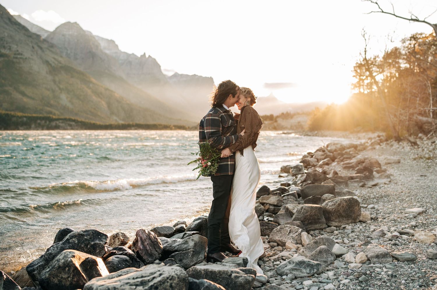 Two people embrace on a rocky lakeshore at sunset with misty mountains and water in the background.
