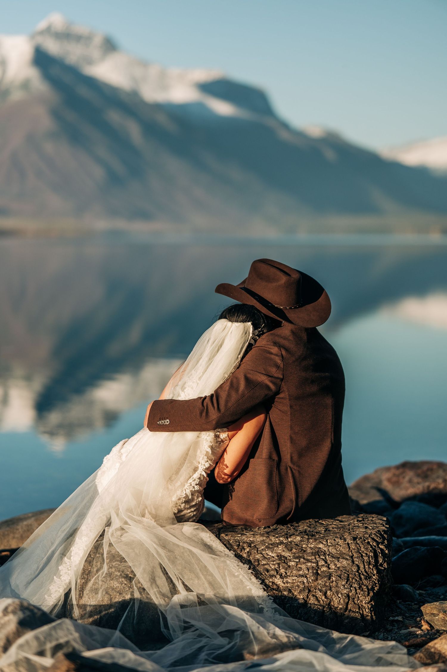 A romantic couple rests on rocks by a serene mountain lake during sunset with a flowing white dress and brown jacket.