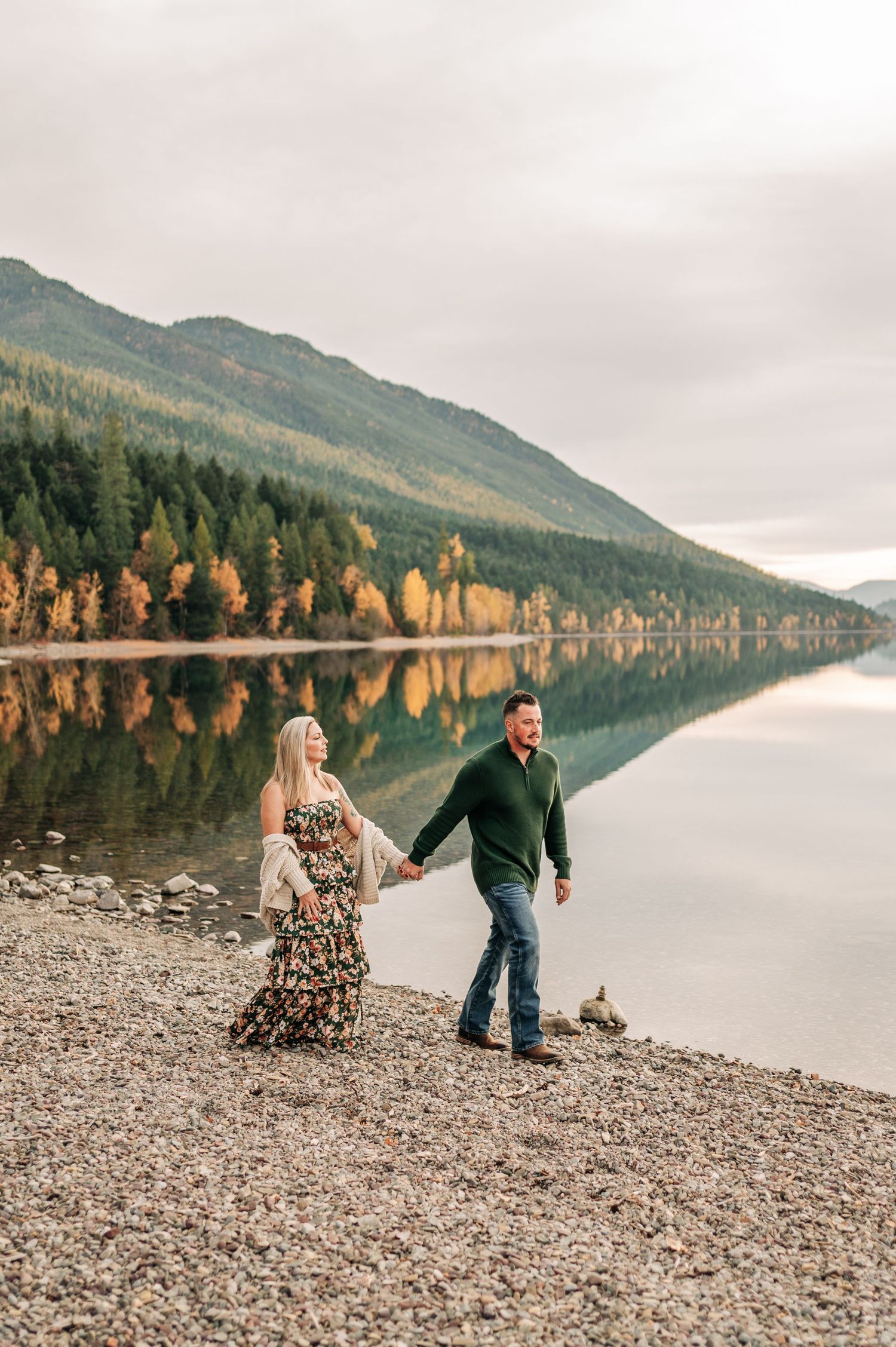 A couple walks hand in hand along a rocky shoreline with mountains and autumn trees reflecting in the calm lake waters.