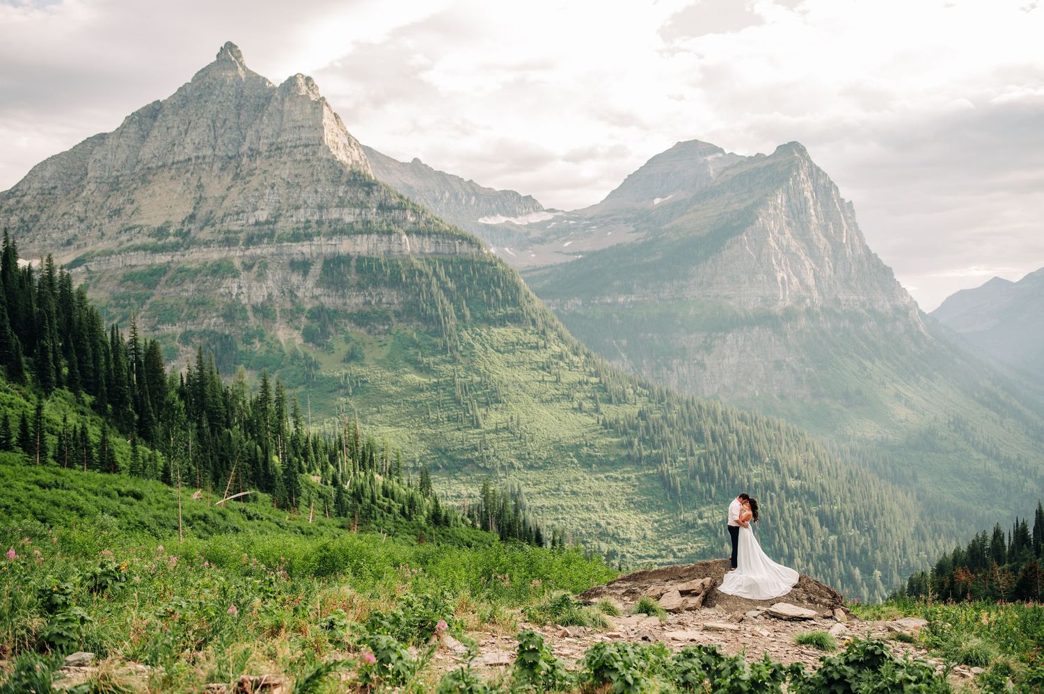 A panoramic mountain landscape view in Glacier National Park with snow-capped peaks and lush green valleys stretches out below.