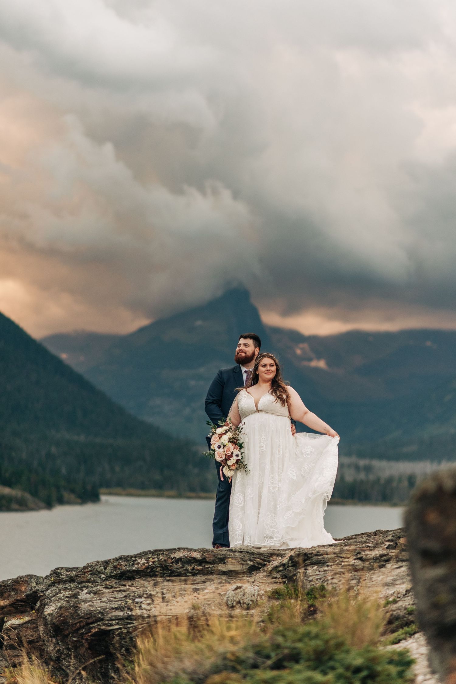 wedding couple standing on a rock in glacier national park with moody skies behind them at sunset