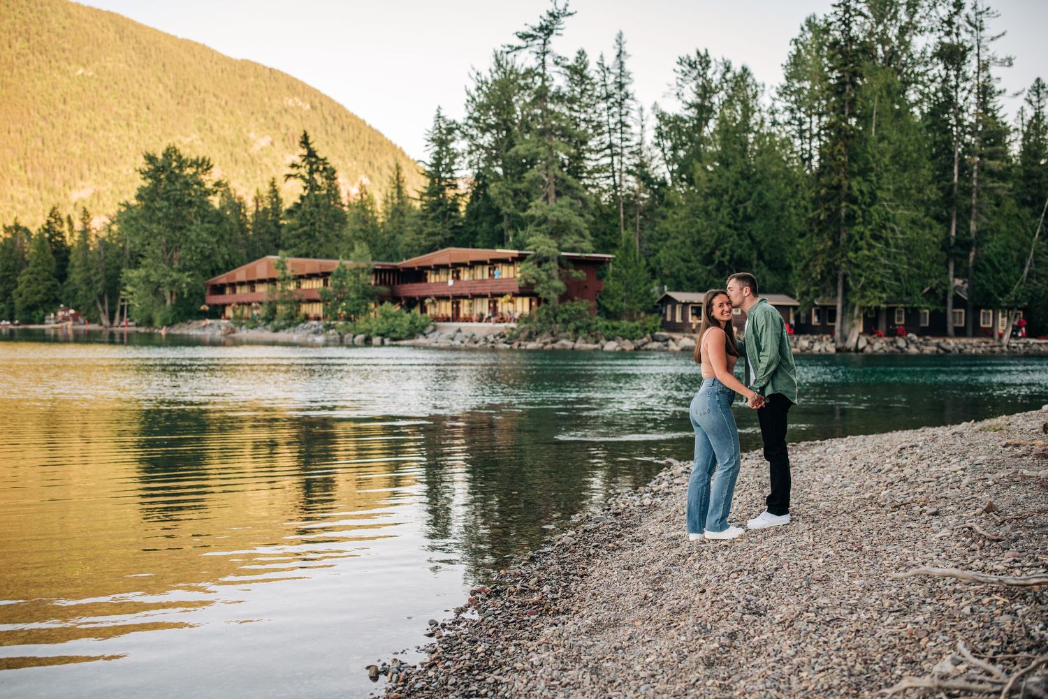 Couple stands on rocky lakeshore at sunset with rustic mountain lodge and evergreen trees in background.