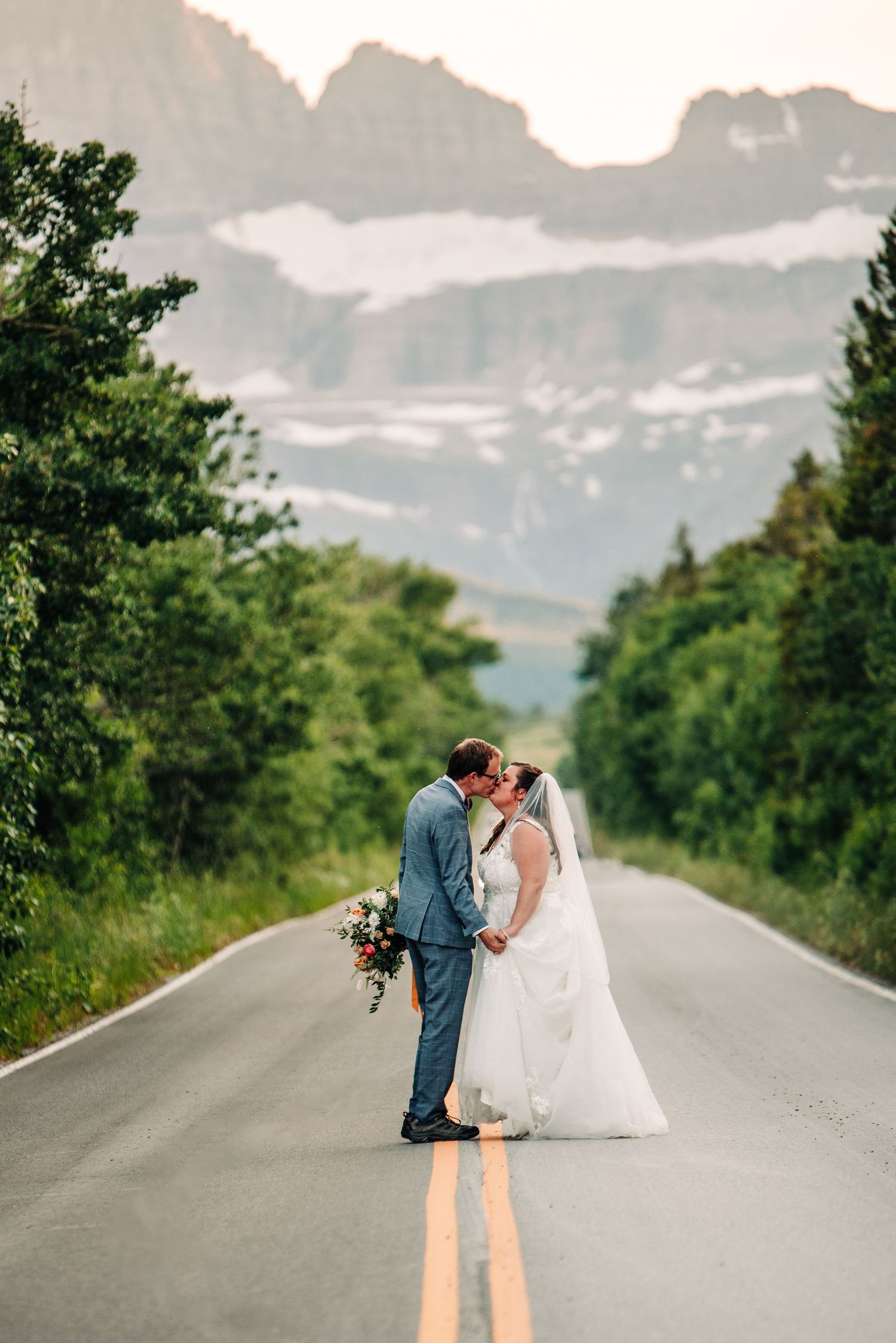 A couple shares a romantic kiss on an empty mountain road with dramatic snow-capped peaks in the background.