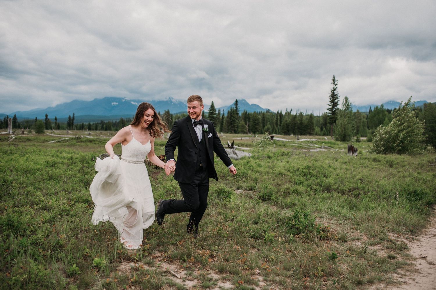 A couple in wedding attire walks joyfully through a scenic mountain meadow with dramatic cloudy skies overhead.