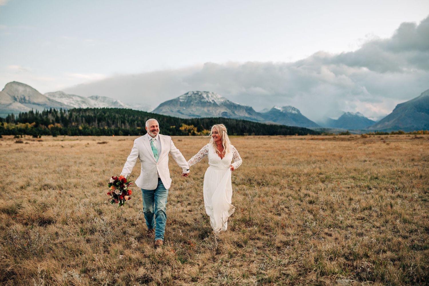 Two people in formal attire walk hand in hand across a golden field with snow-capped mountains in the background.