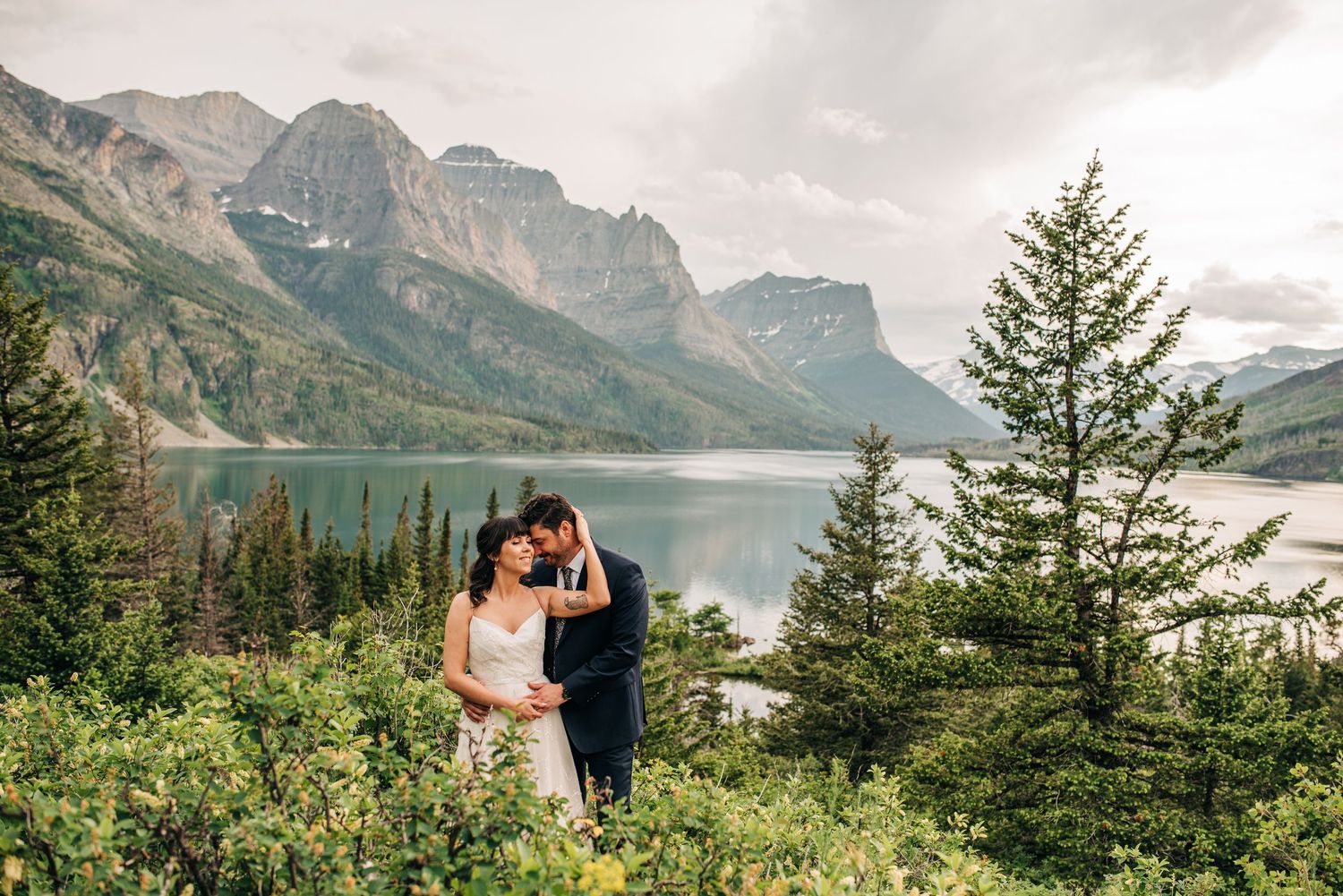 Couple embracing at scenic Glacier National Park overlook with turquoise lake and mountain peaks in background.