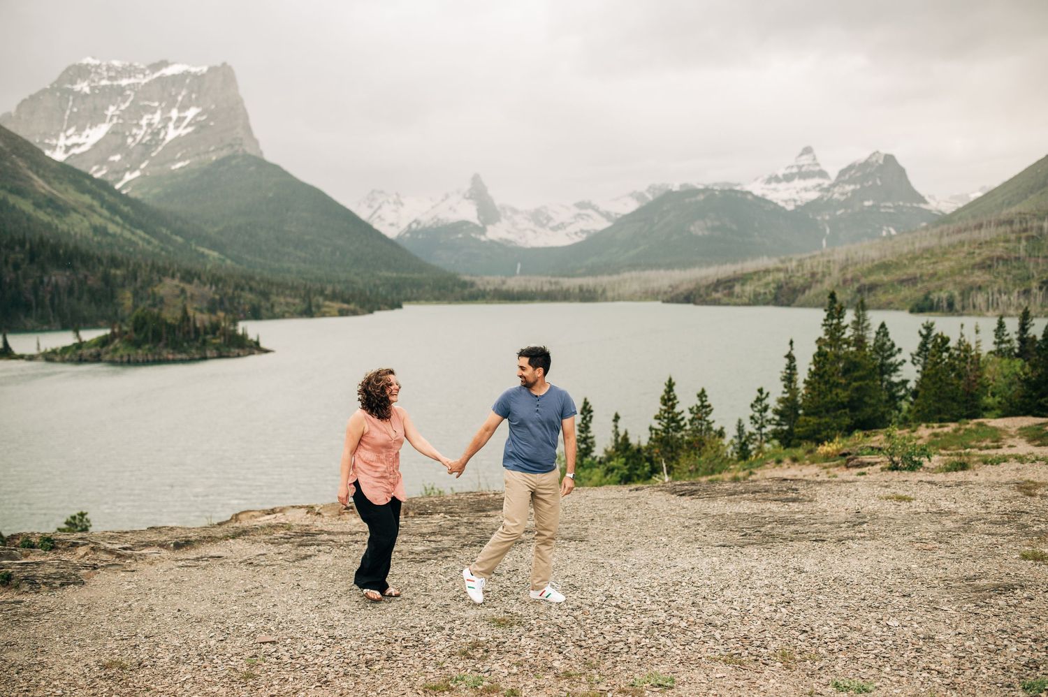 Two people enjoy a scenic mountain view overlooking a pristine lake surrounded by evergreen trees and snow-capped peaks.