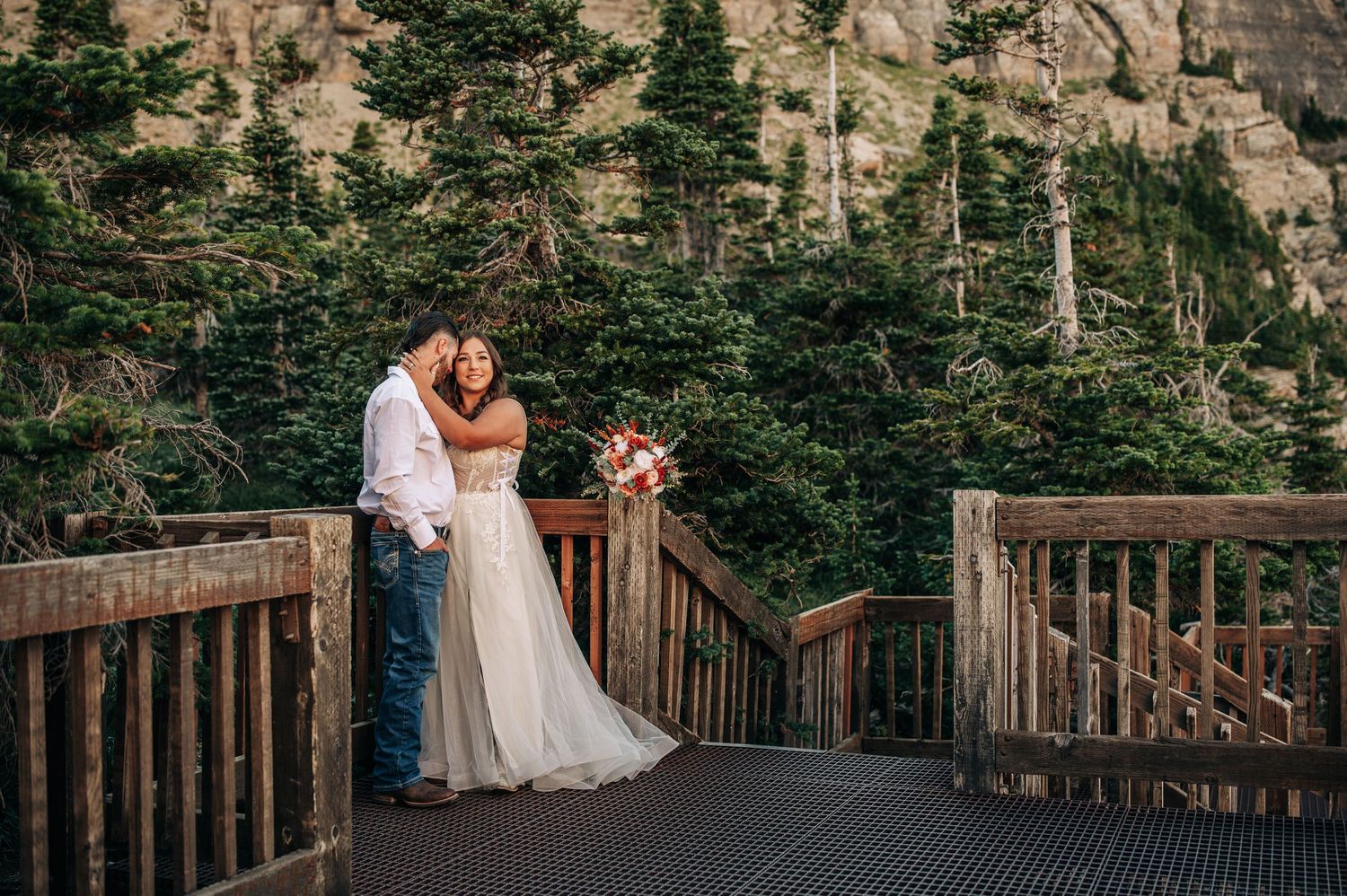 A couple standing on a wooden boardwalk surrounded by evergreen trees and mountain cliffs in a scenic outdoor setting.