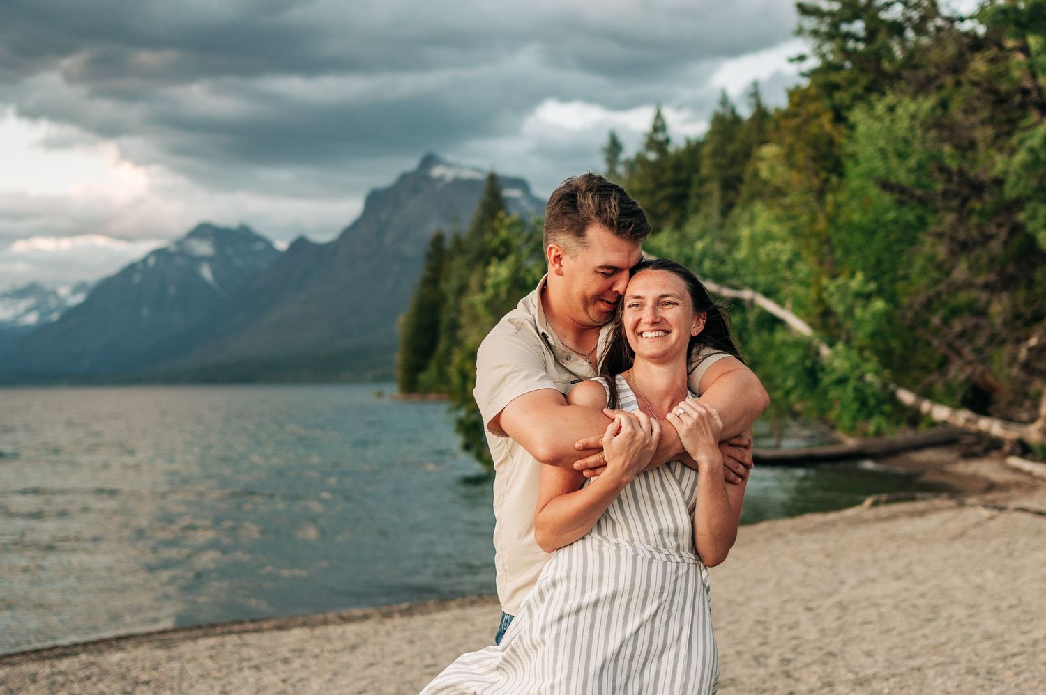 A loving couple embraces on a beach shore with dramatic mountains and forest in the background at Grand Teton National Park.