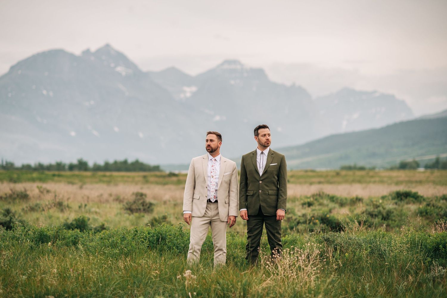 Two people in formal suits stand in a meadow with majestic mountain peaks rising in the misty background.