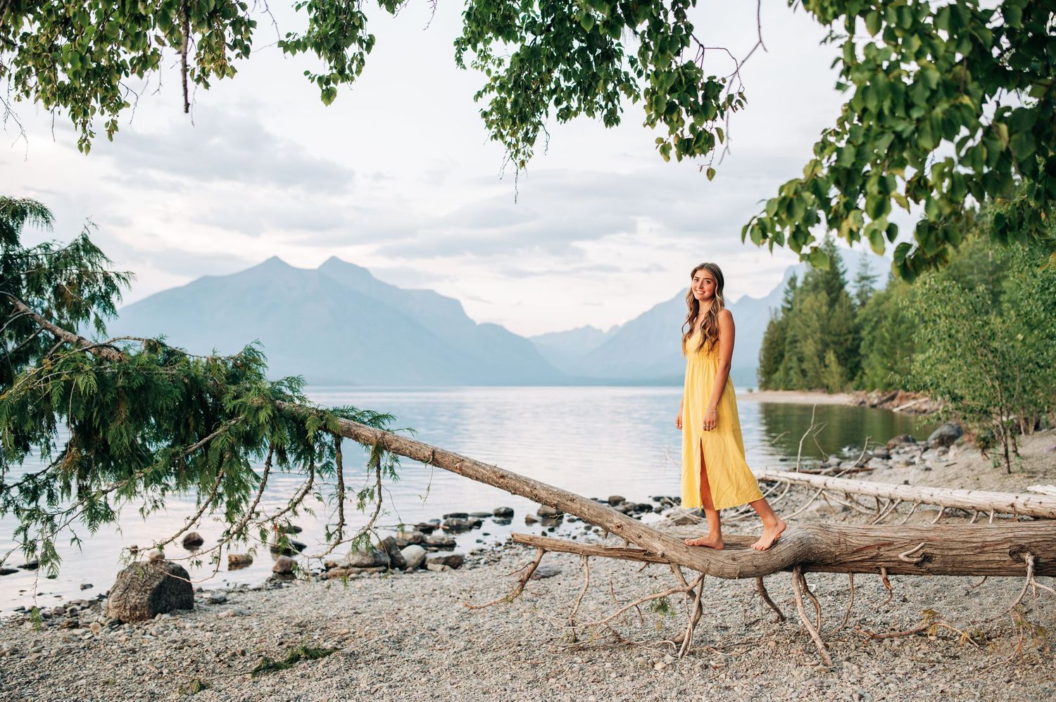 Person in yellow dress stands on driftwood log at scenic mountain lake shore with evergreen trees and cloudy sky.
