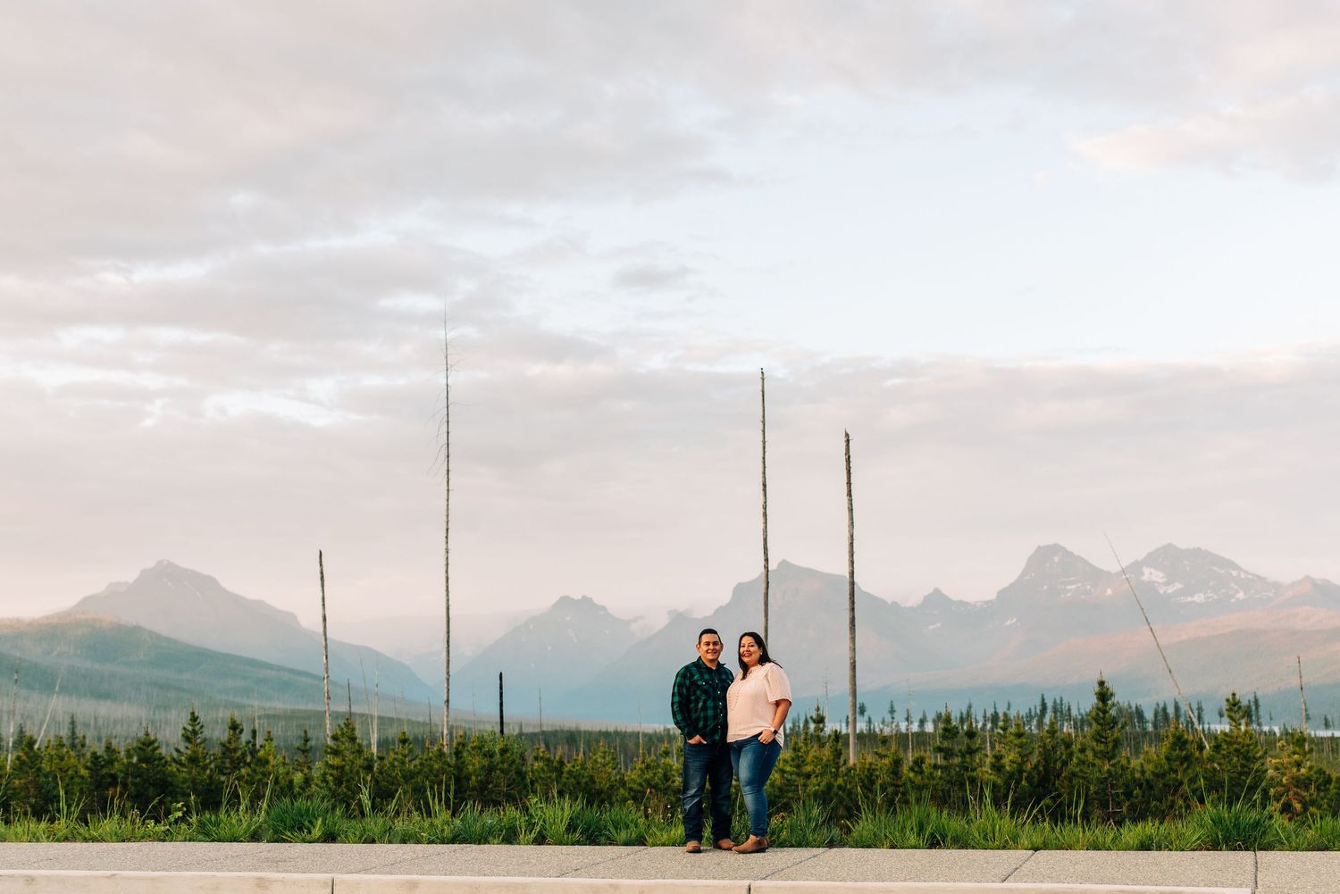 Two people stand together on a path with majestic mountain peaks and green fields stretching into the distance.