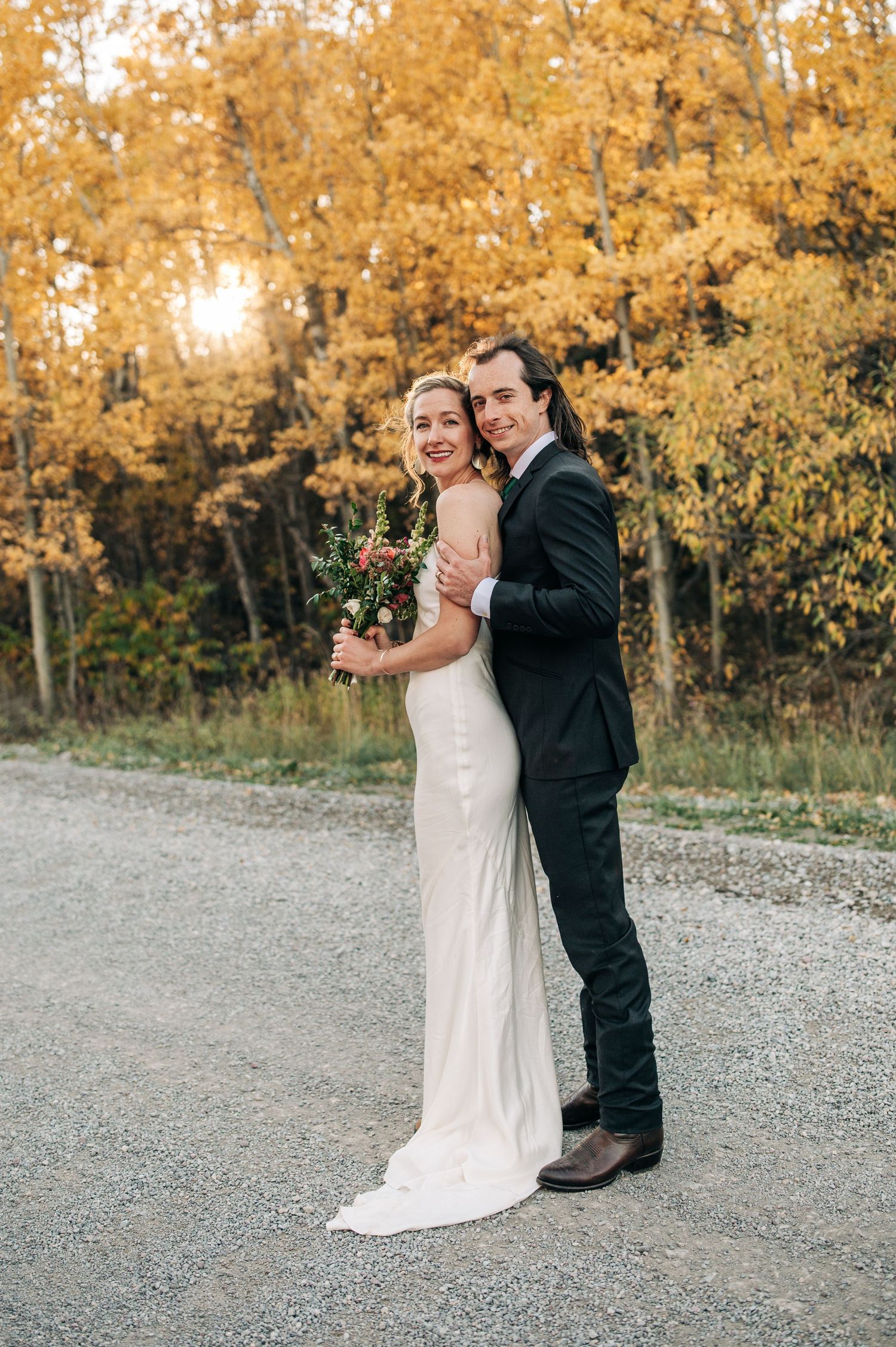 A couple in wedding attire poses on a gravel path surrounded by golden autumn trees at sunset.