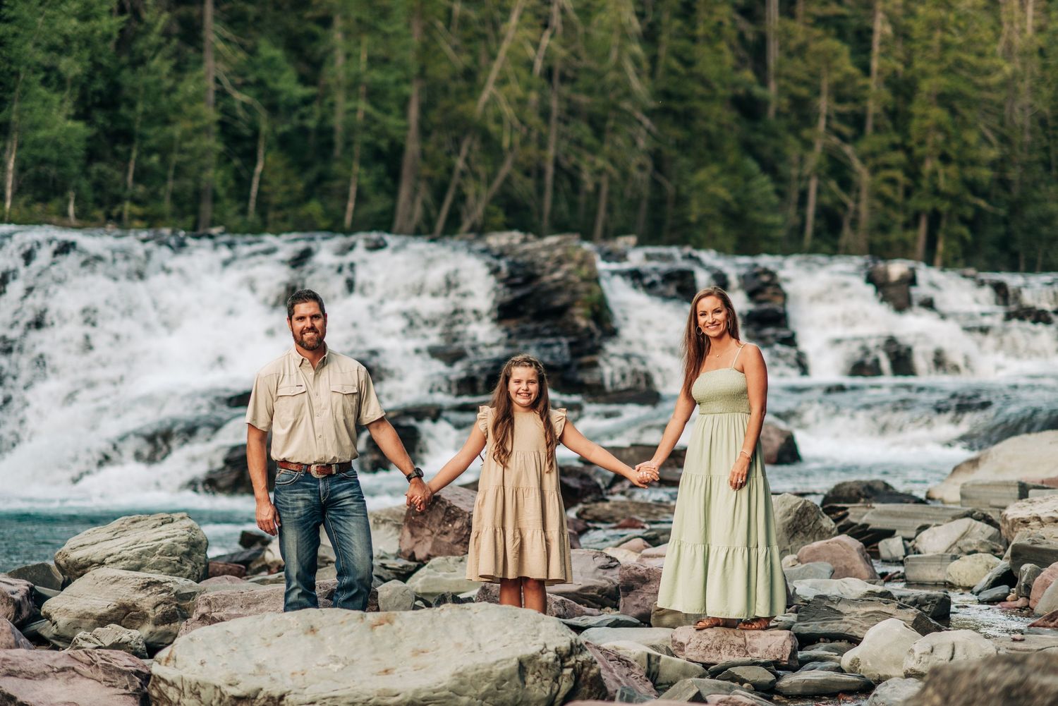 Three people standing on rocks near cascading waterfalls in a forest setting wearing light colored casual attire.