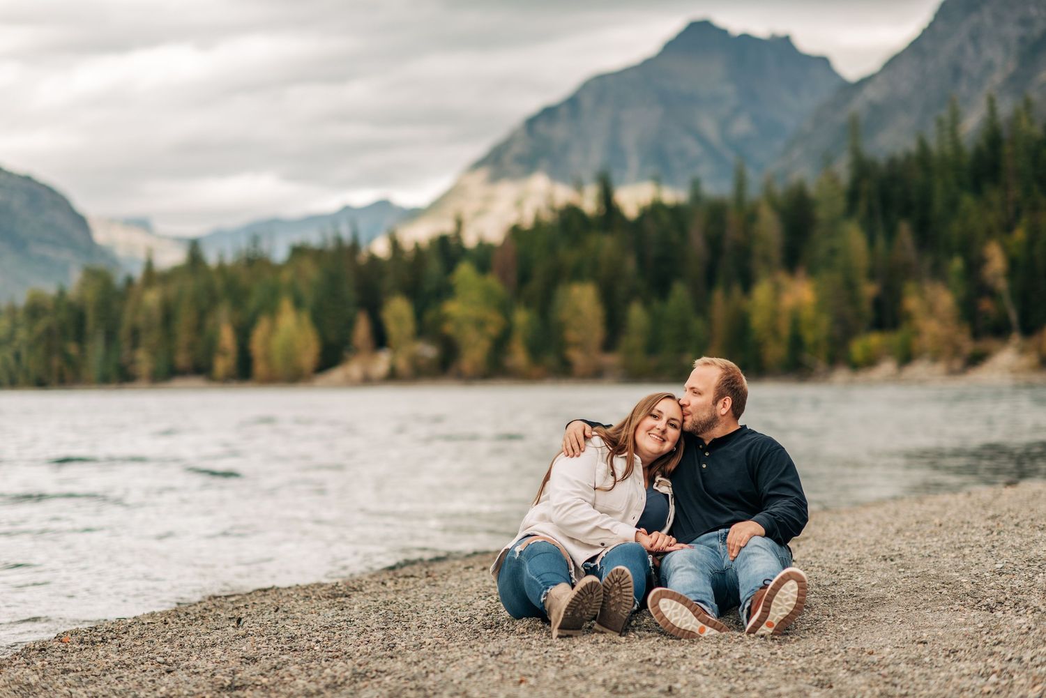 Two people sit together on a rocky beach beside a mountain lake surrounded by autumn trees and rugged peaks.