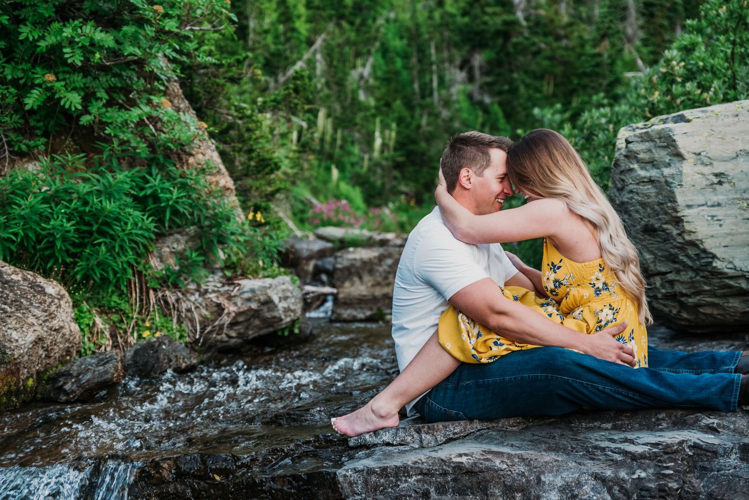 Two people in casual attire sit intimately by a rocky stream in a lush forest setting.