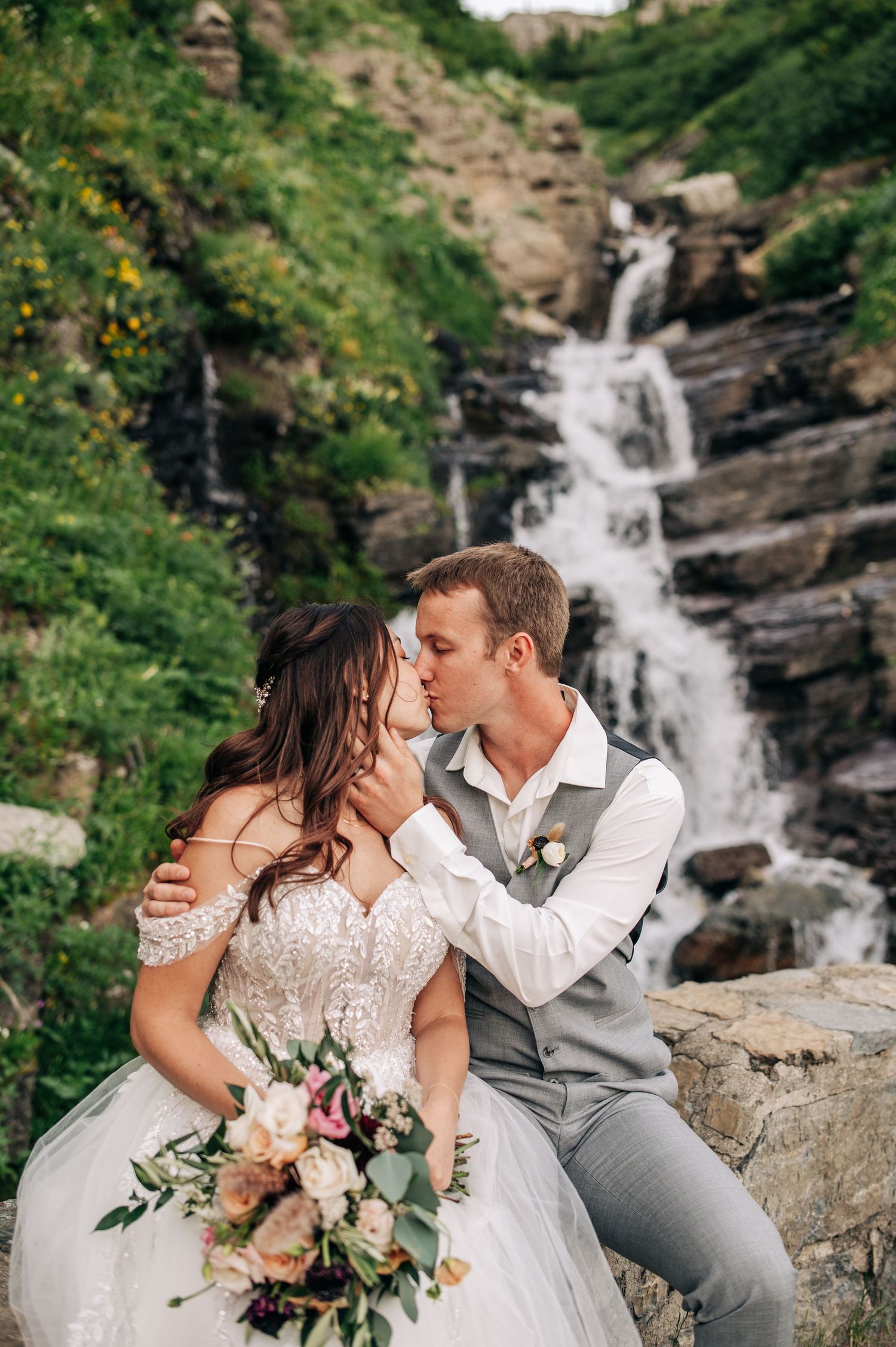 A romantic couple shares a tender moment near a cascading waterfall, with the bride holding a beautiful floral bouquet.