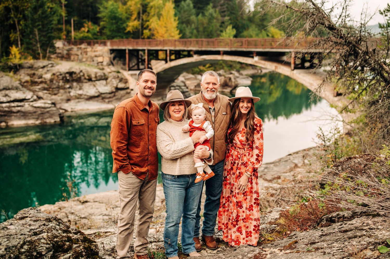 A group of people stand together on a rocky riverbank with a stone bridge and turquoise water in the background.