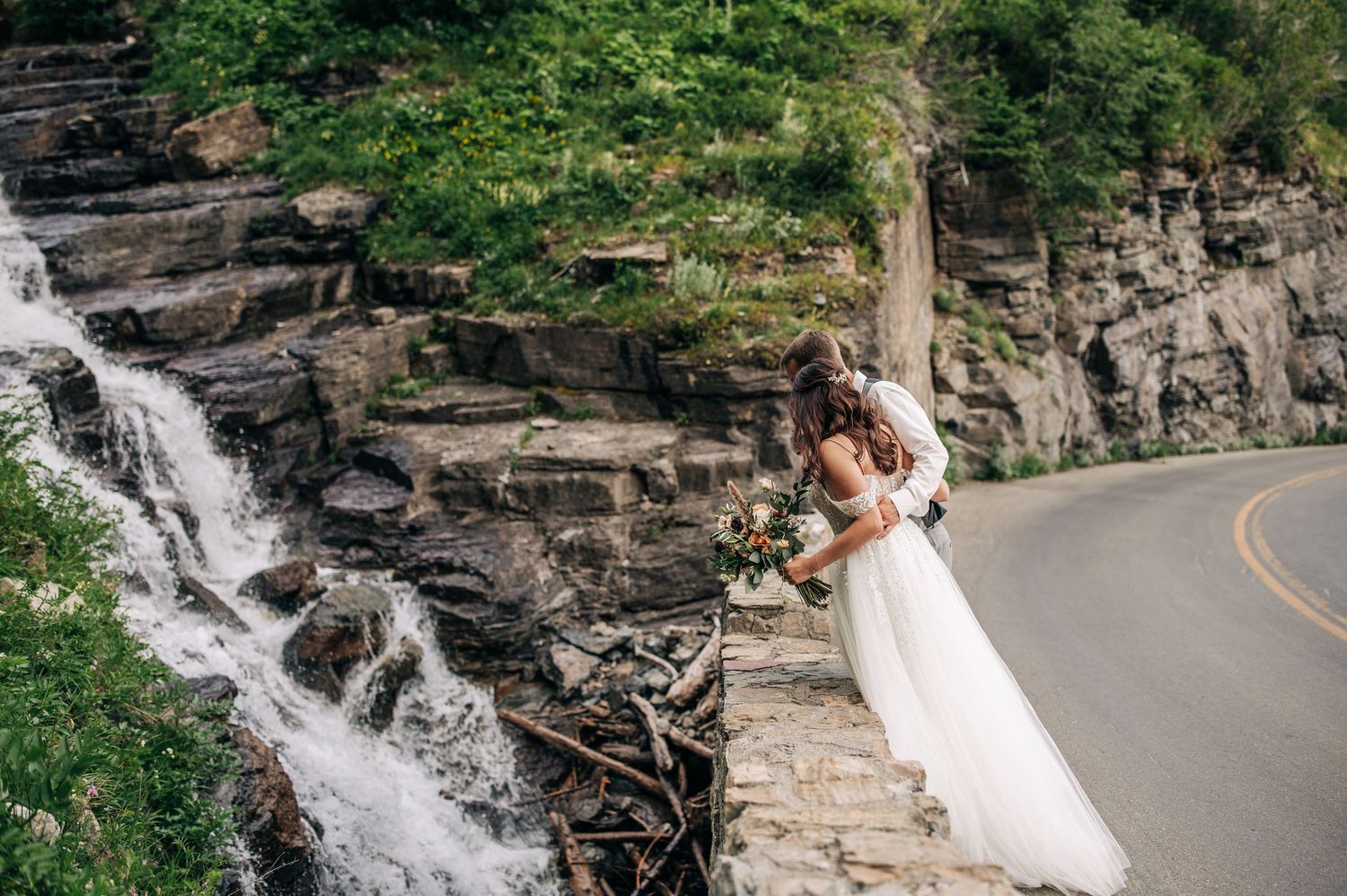 Couple embracing beside cascading waterfall on winding mountain road with stone retaining wall.