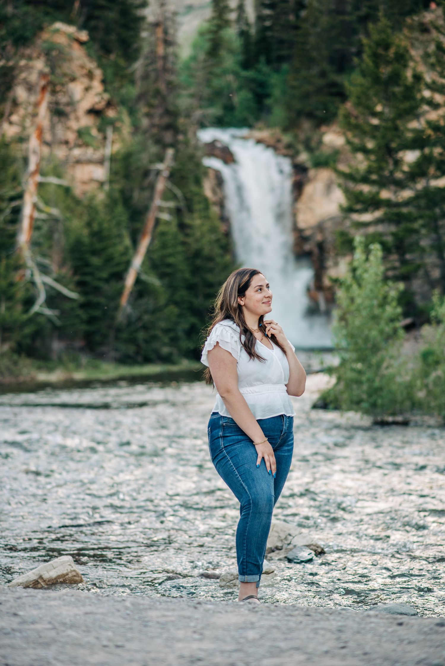 A person in jeans poses playfully on a rocky riverbank with a scenic waterfall cascading in the forest background.