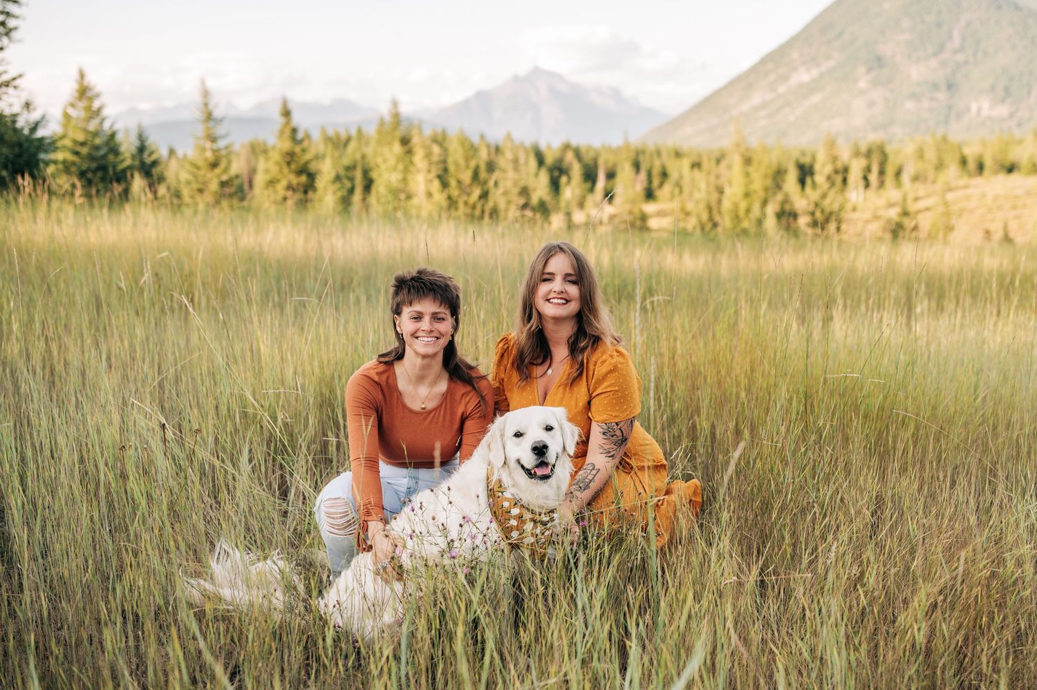 A happy white dog sits in a golden meadow with mountain views and evergreen trees in the background.