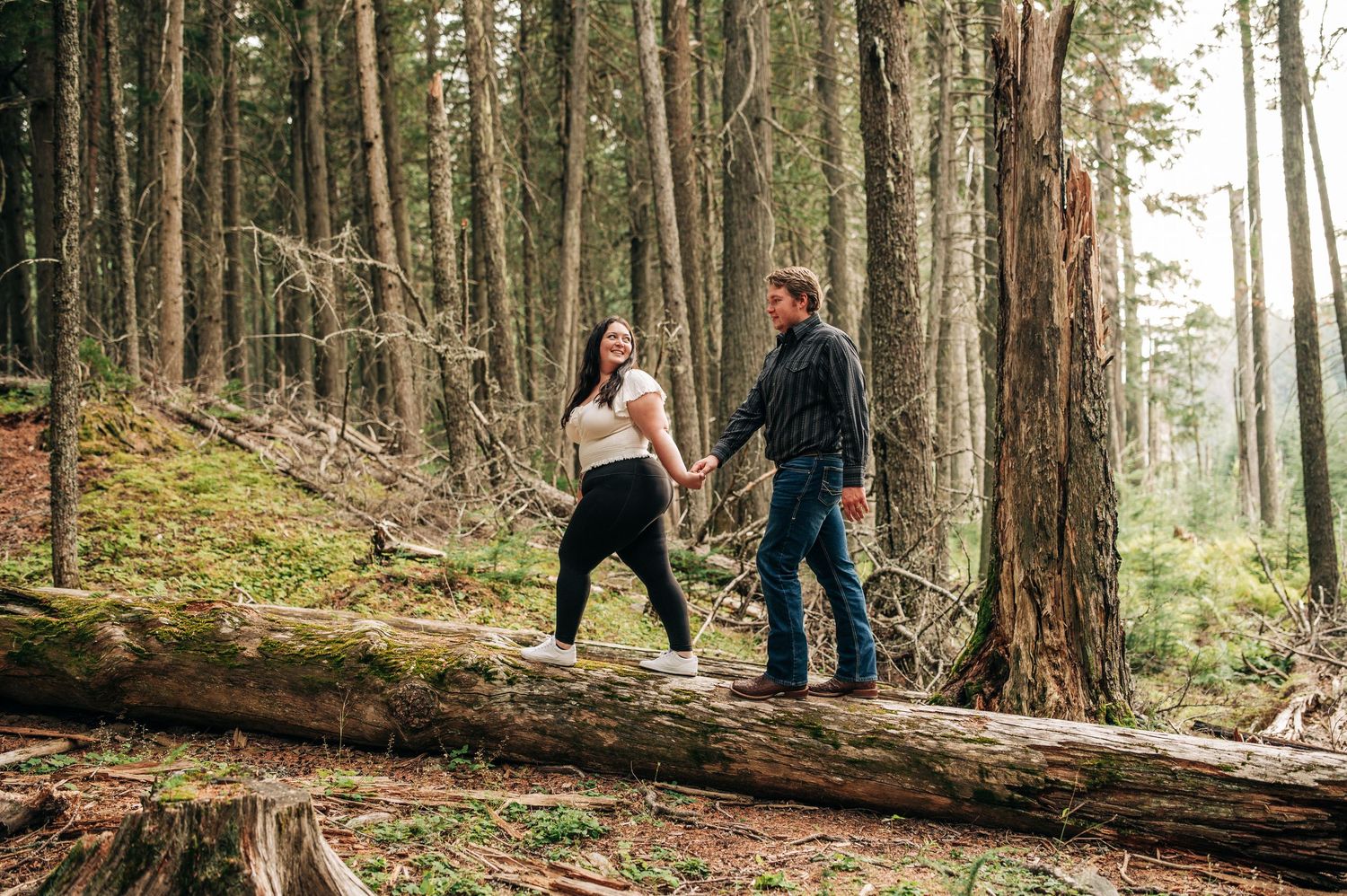 Two people balance carefully while walking across a large fallen log in a misty evergreen forest.