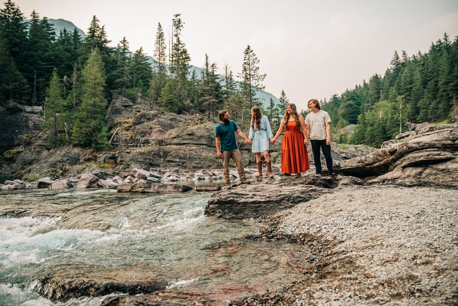 Group standing on rocky shore near evergreen forest beside rushing mountain river with cloudy skies.