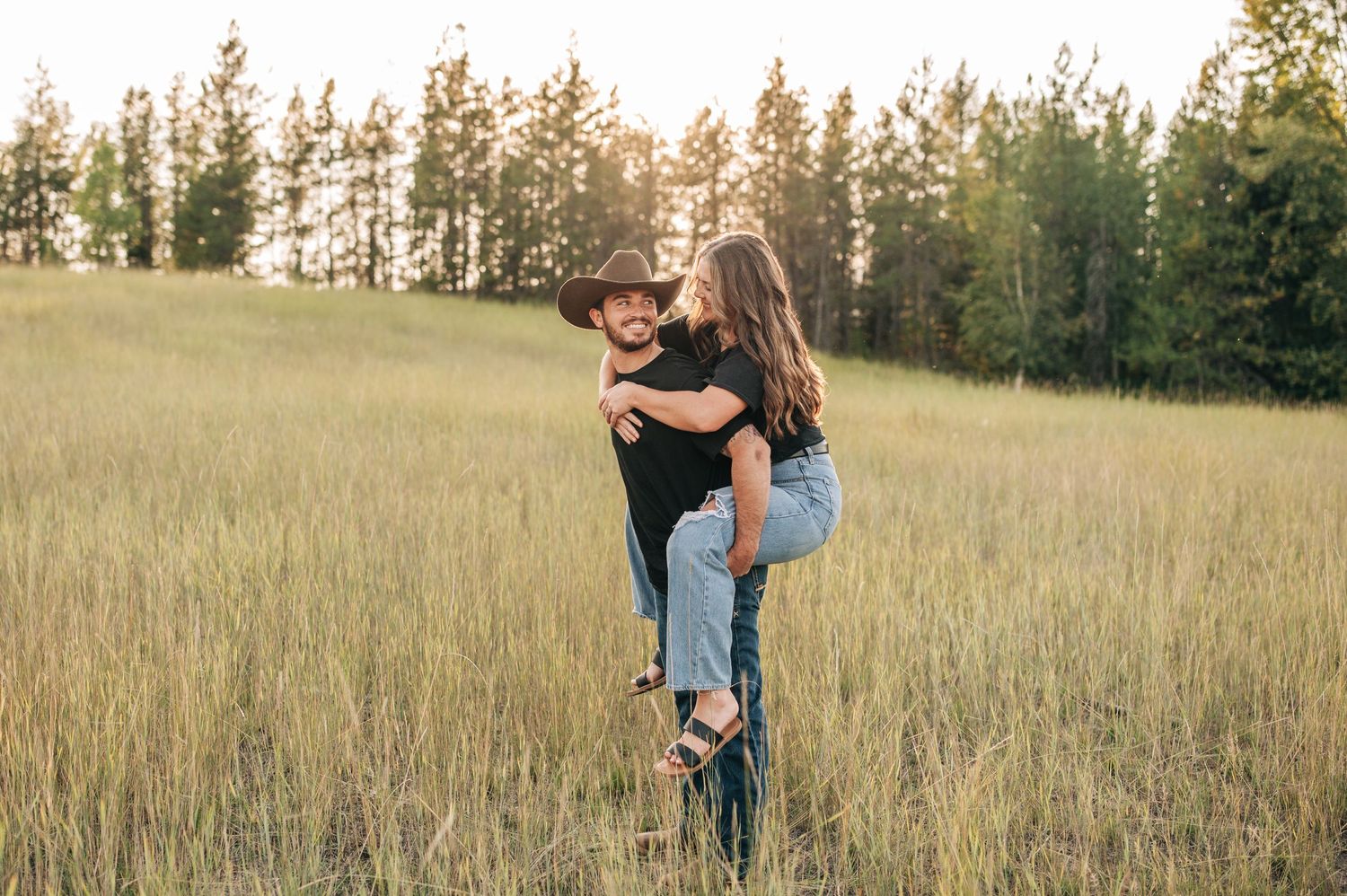 A couple embraces in a golden field at sunset with evergreen trees in the background.