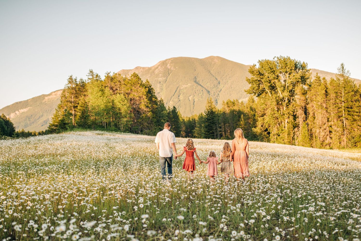 A group walks through a wildflower meadow with golden trees and a mountain peak in the background at sunset.