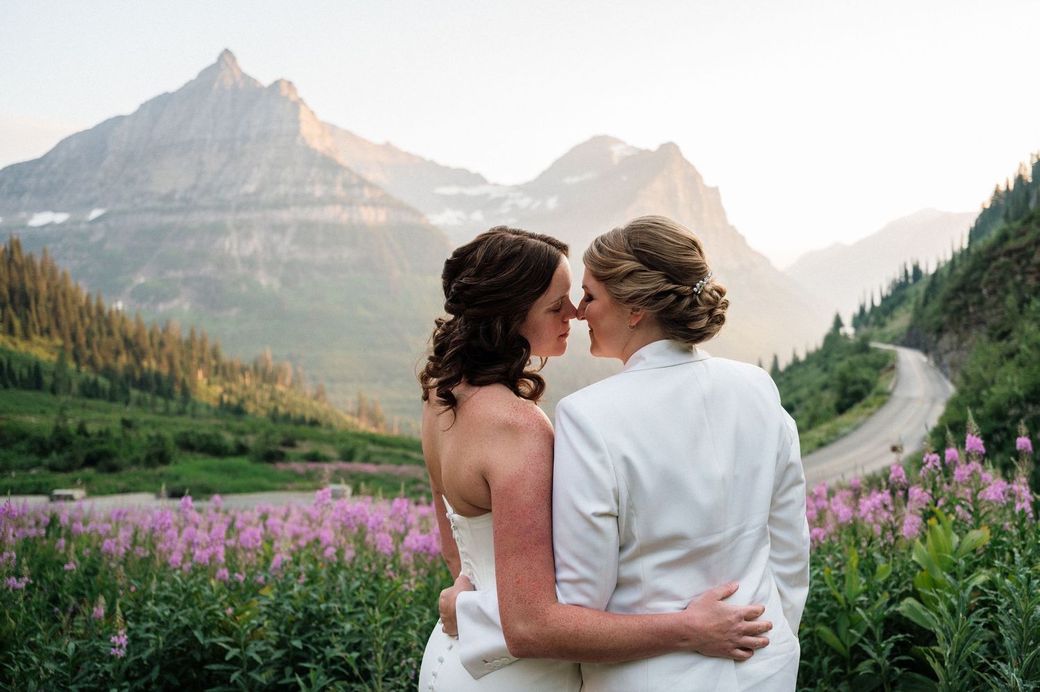 Two brides share a romantic moment in a mountain meadow with wildflowers and scenic road backdrop.