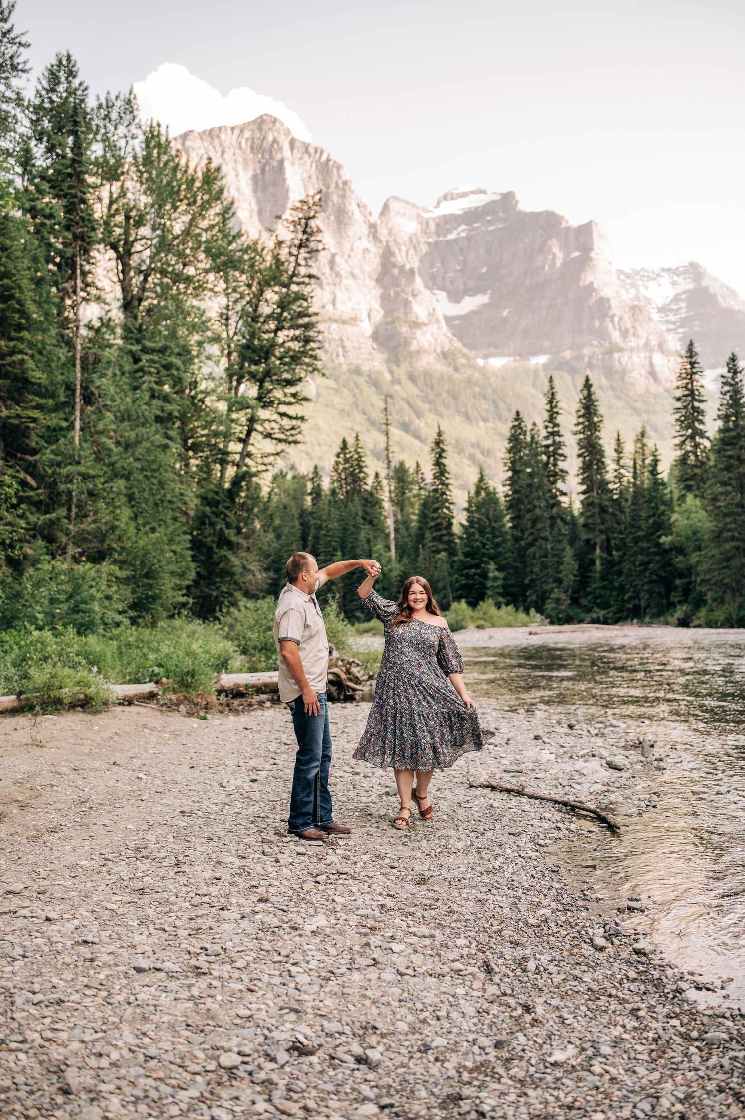 People gather on a rocky shoreline surrounded by evergreen trees and majestic mountain peaks in a serene wilderness setting.