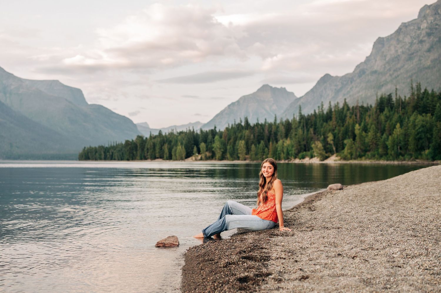 Person sitting on rocky shore of mountain lake surrounded by pine forests and towering peaks at sunset.