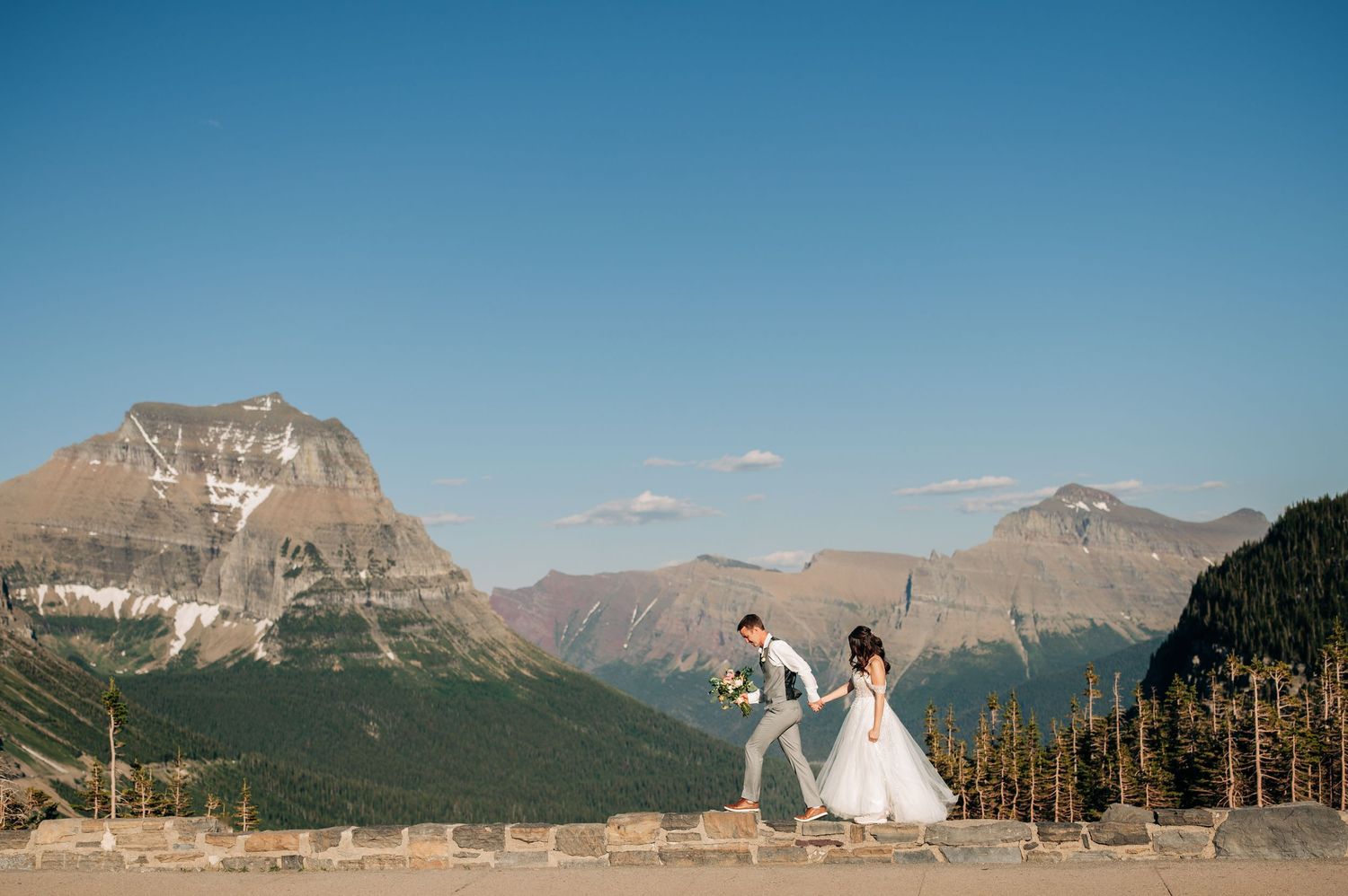 Two people celebrating against a stunning mountain backdrop in Glacier National Park on a clear sunny day.