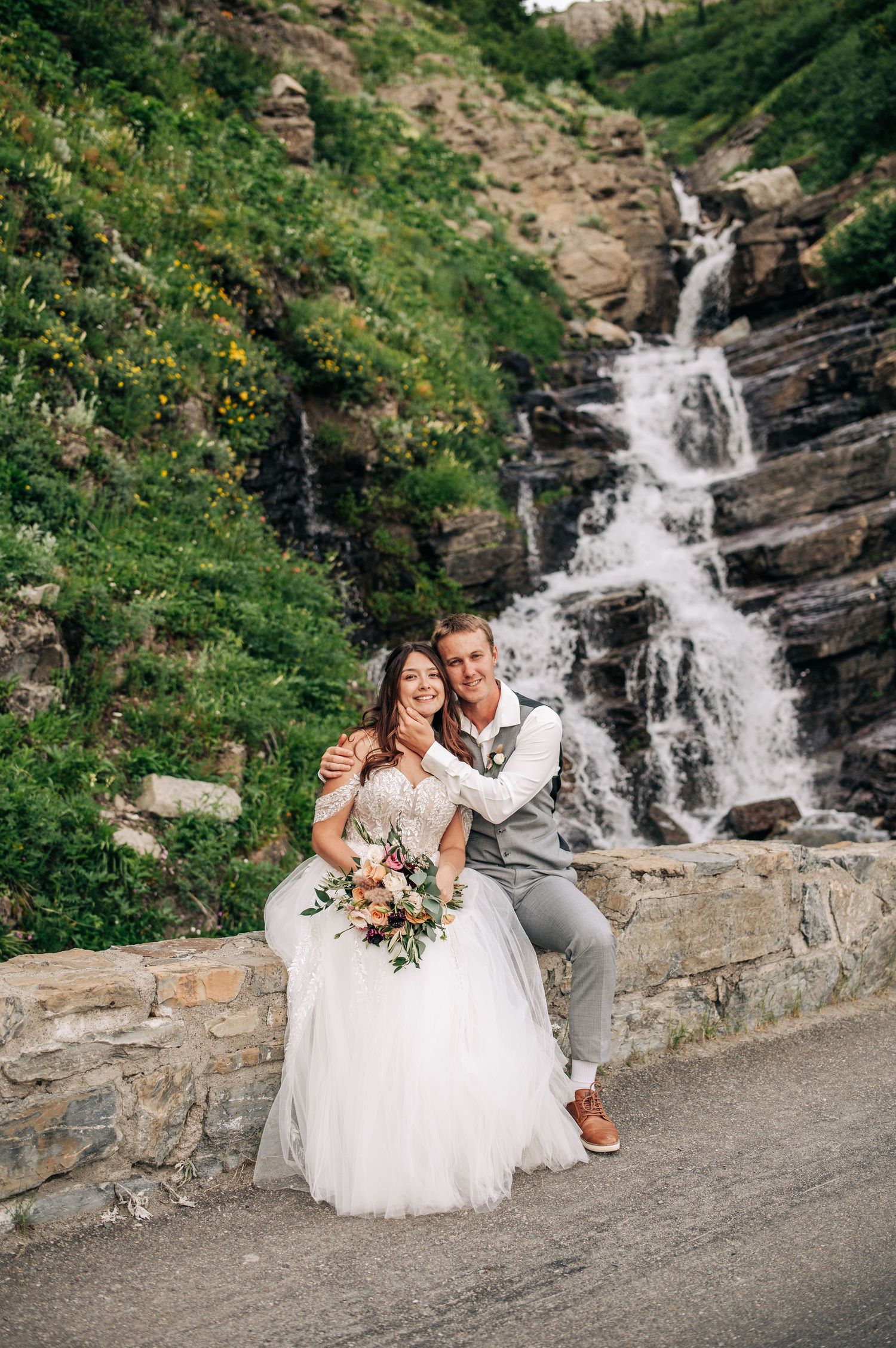 A wedding couple sits on a stone wall with a cascading mountain waterfall and lush greenery in the background.
