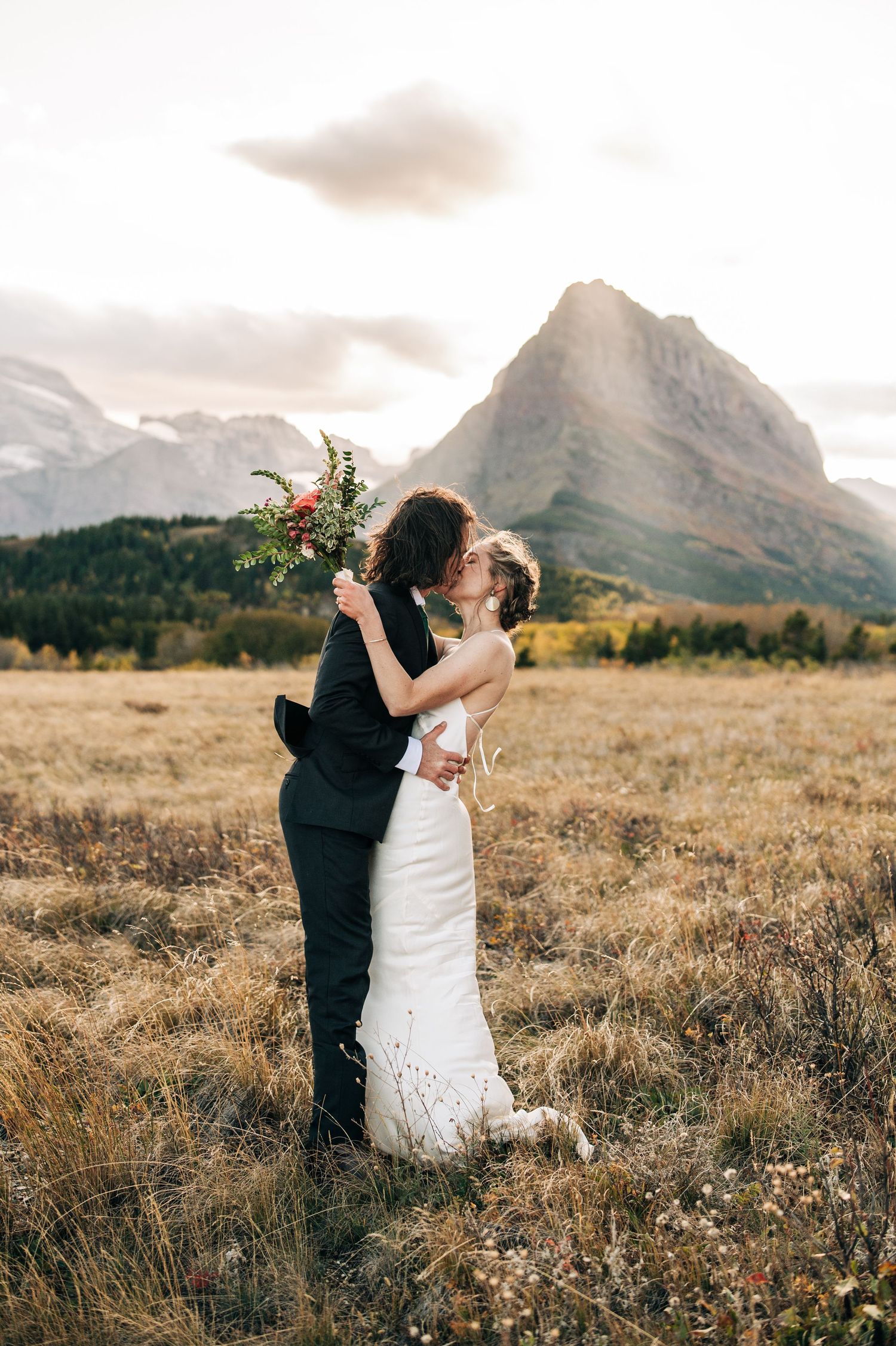 Two figures share a romantic moment in a golden field with misty mountains looming in the background at sunset.