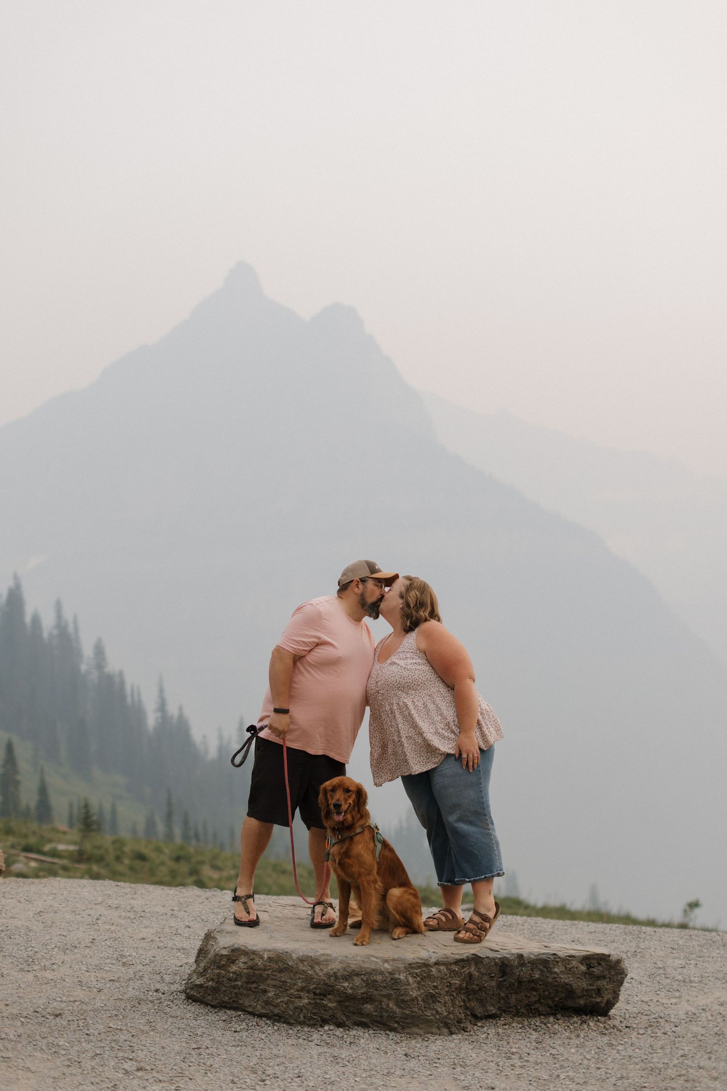 Couple kissing on a rock with their golden dog, misty mountain backdrop, outdoor engagement photo.
