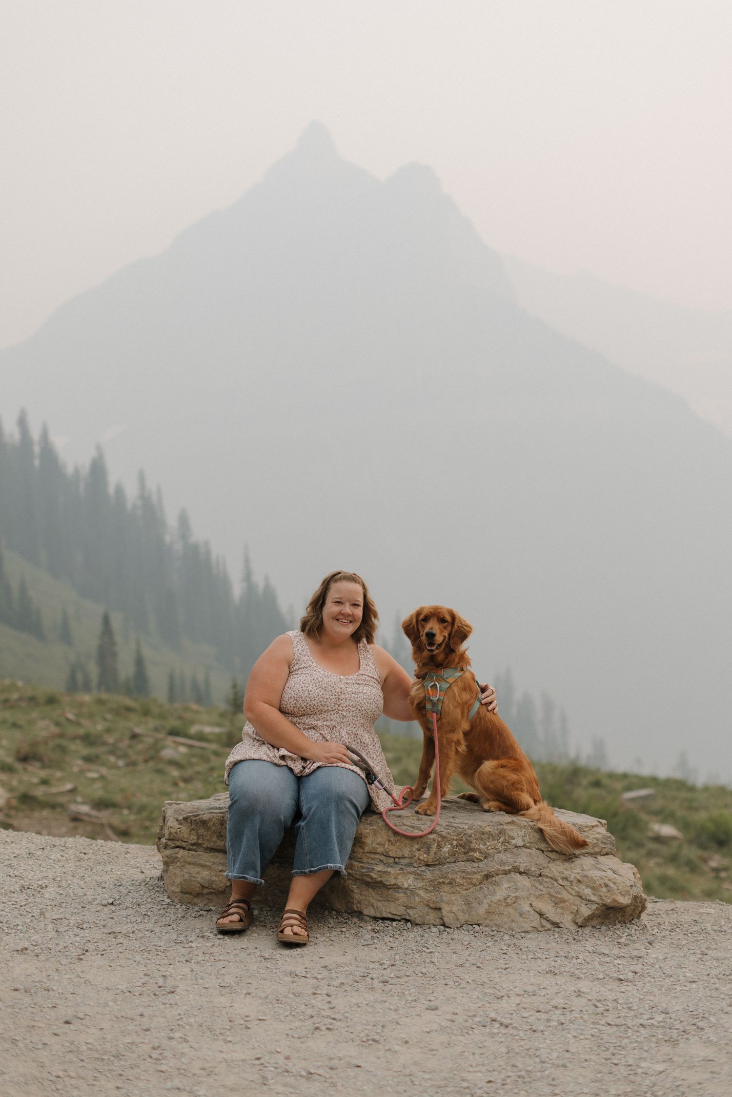 Woman sits on a rock with her golden retriever dog, smiling before a misty mountain peak and evergreen forest backdrop.